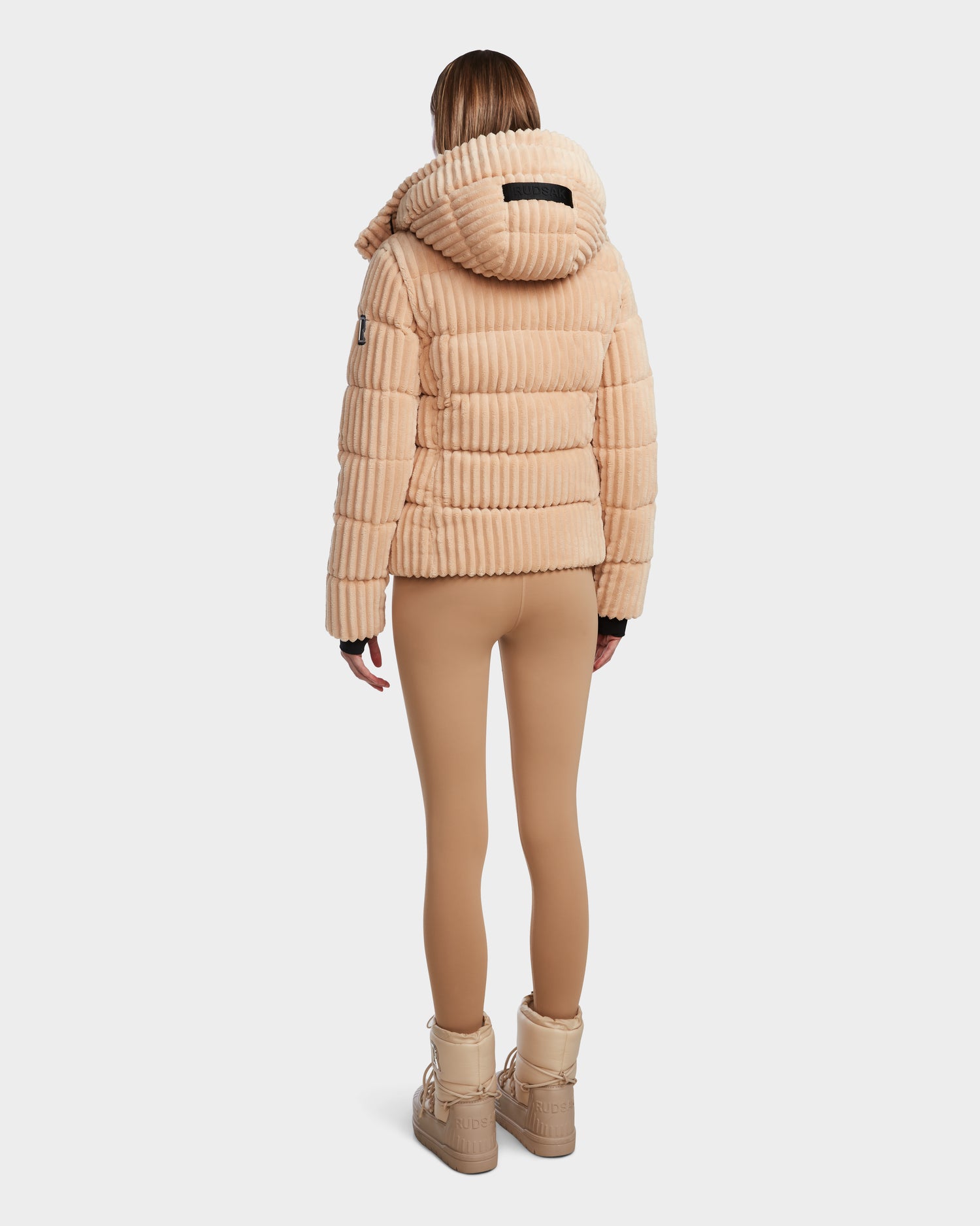 A person faces away, wearing the MIRRY FF - L124507 Iceland beige velvet corduroy puffer with hood by Rudsak, tan leggings, and beige boots against a plain white background.