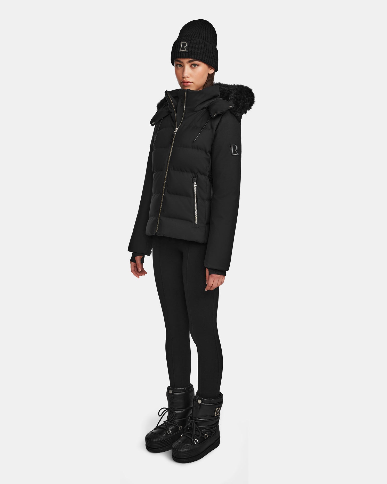 A woman wearing the Rudsak MIRRY M - L124533 Black quilted down coat with fur-trimmed hood, plus a black knit beanie, leggings, and winter boots stands against a plain white background.
