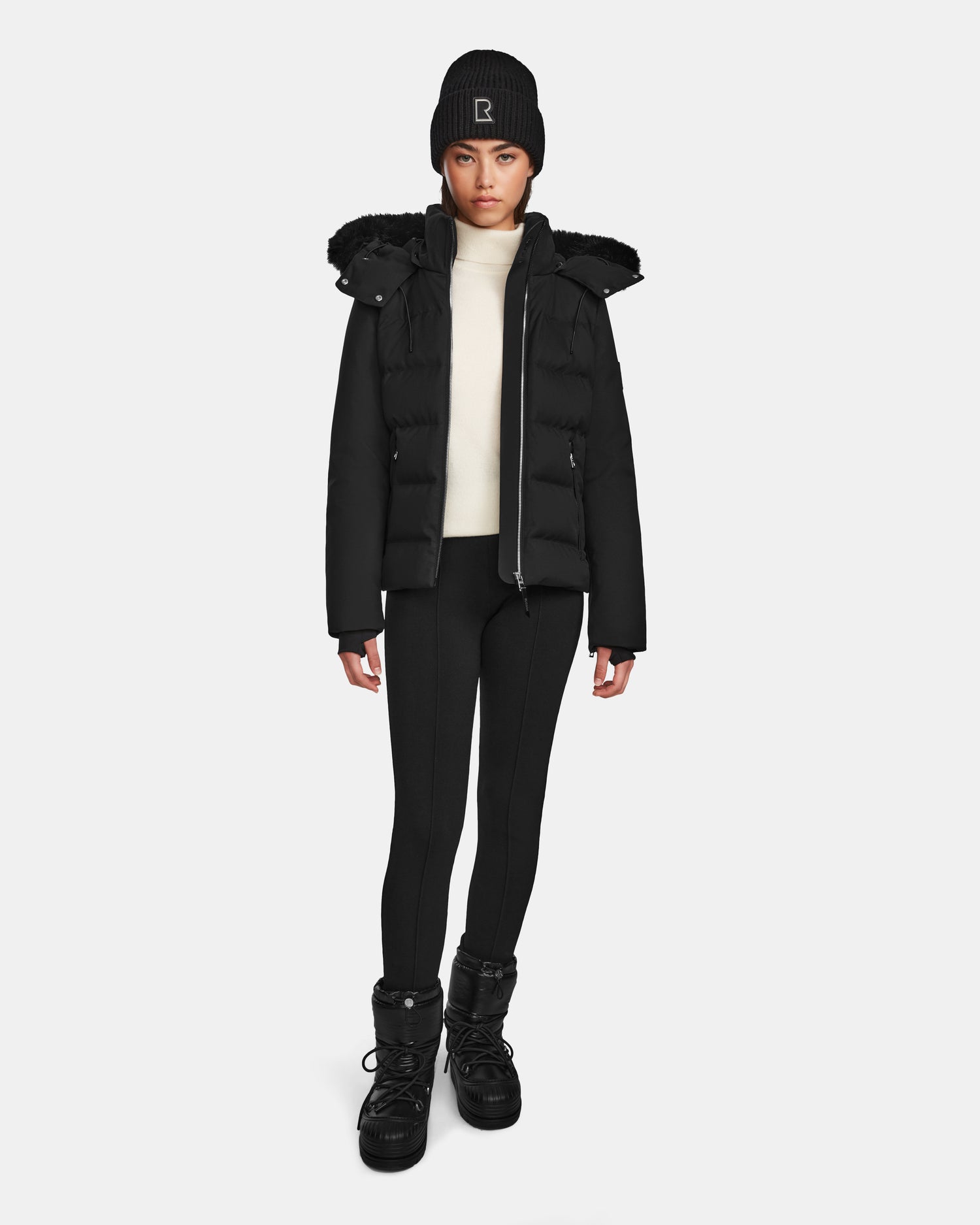 A person stands facing forward wearing the Rudsak MIRRY M - L124533 Black women's quilted down coat with a fur-trimmed hood, paired with a black beanie, white turtleneck, black leggings, and winter boots against a light background.