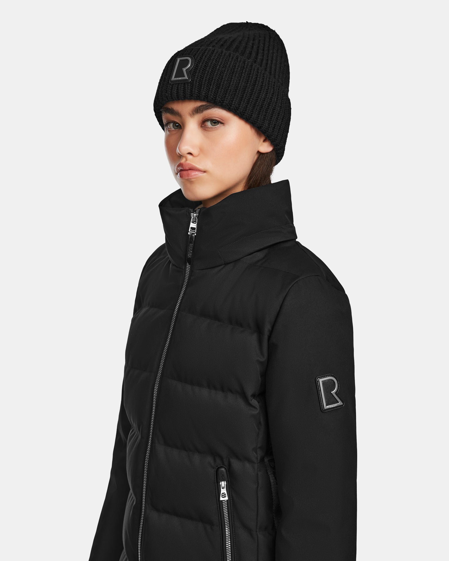 A person in the Rudsak MIRRY M - L124533 Black beanie and zip-up puffer jacket with embroidered R logos, styled like a women's quilted down coat made from RDS-certified down, stands against a plain background.