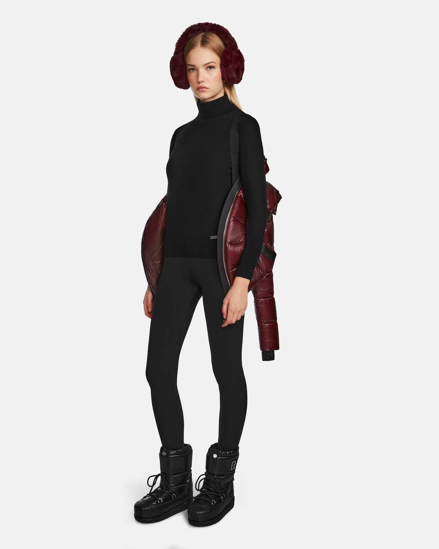 A woman faces forward against a plain white background, wearing black leggings, a black turtleneck, black snow boots, red earmuffs, and the Rudsak MIRRY S - L123545 Wine womens quilted down coat draped off her shoulders.