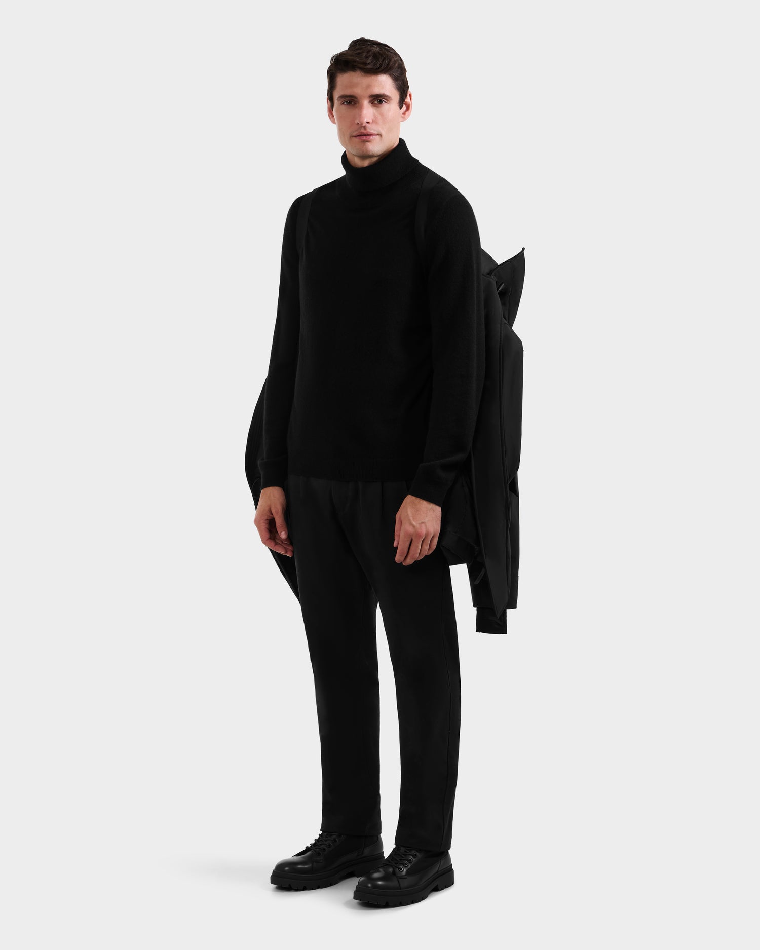 A man faces forward against a plain light gray background, wearing the Rudsak MITCHELL - M125513 Black turtleneck sweater, black pants, and shoes, with a winter parka draped over his left shoulder.
