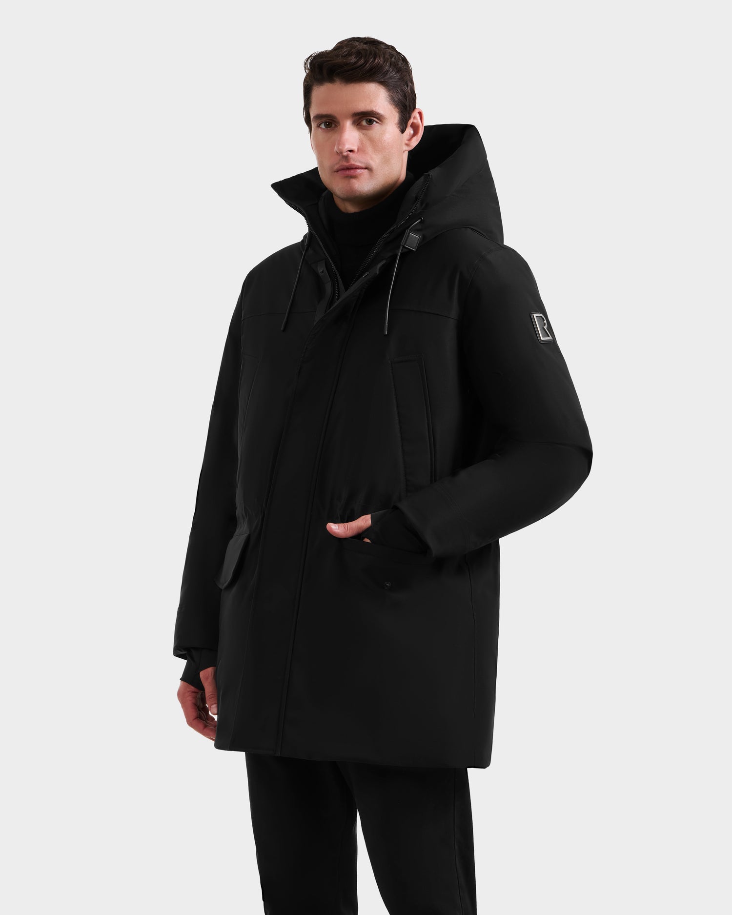 A man in the Rudsak MITCHELL - M125513 Black long men’s down parka with high collar and hood stands against a white background, one hand by his side, the other near the zipper, looking forward.