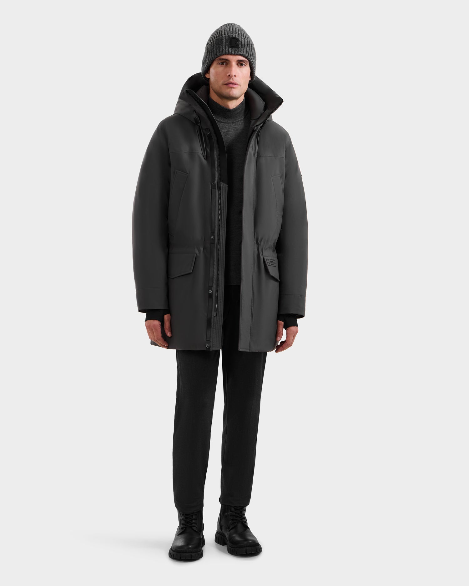 Against a plain white background, a man faces forward wearing the Rudsak MITCHELL - M125513 Grey down winter coat, paired with black pants, boots, a turtleneck sweater, and a gray knit beanie.