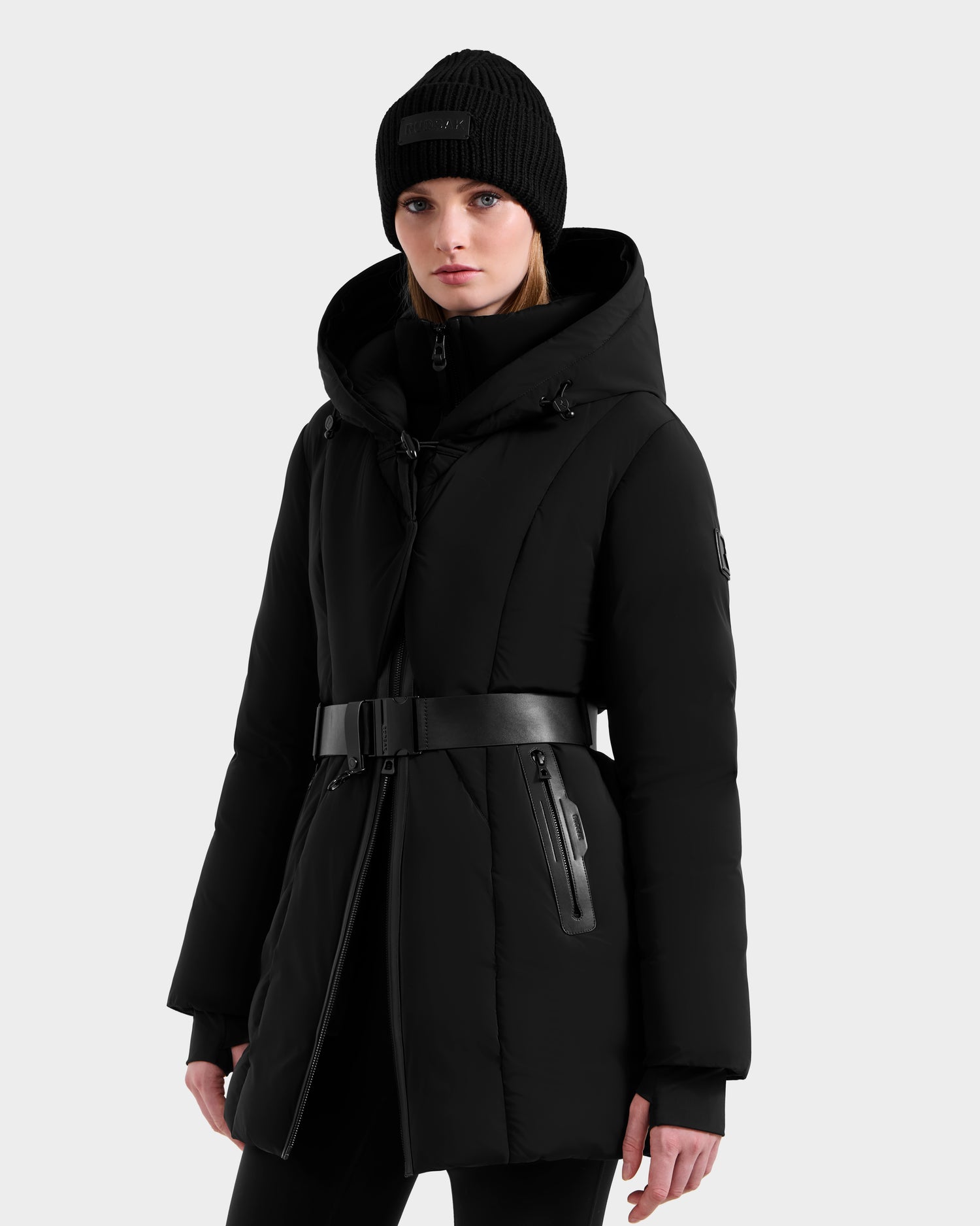 A woman in the Rudsak MODA - 8125704 Black/Natural hooded down parka with a belted waist and a black knit beanie stands against a light gray background.