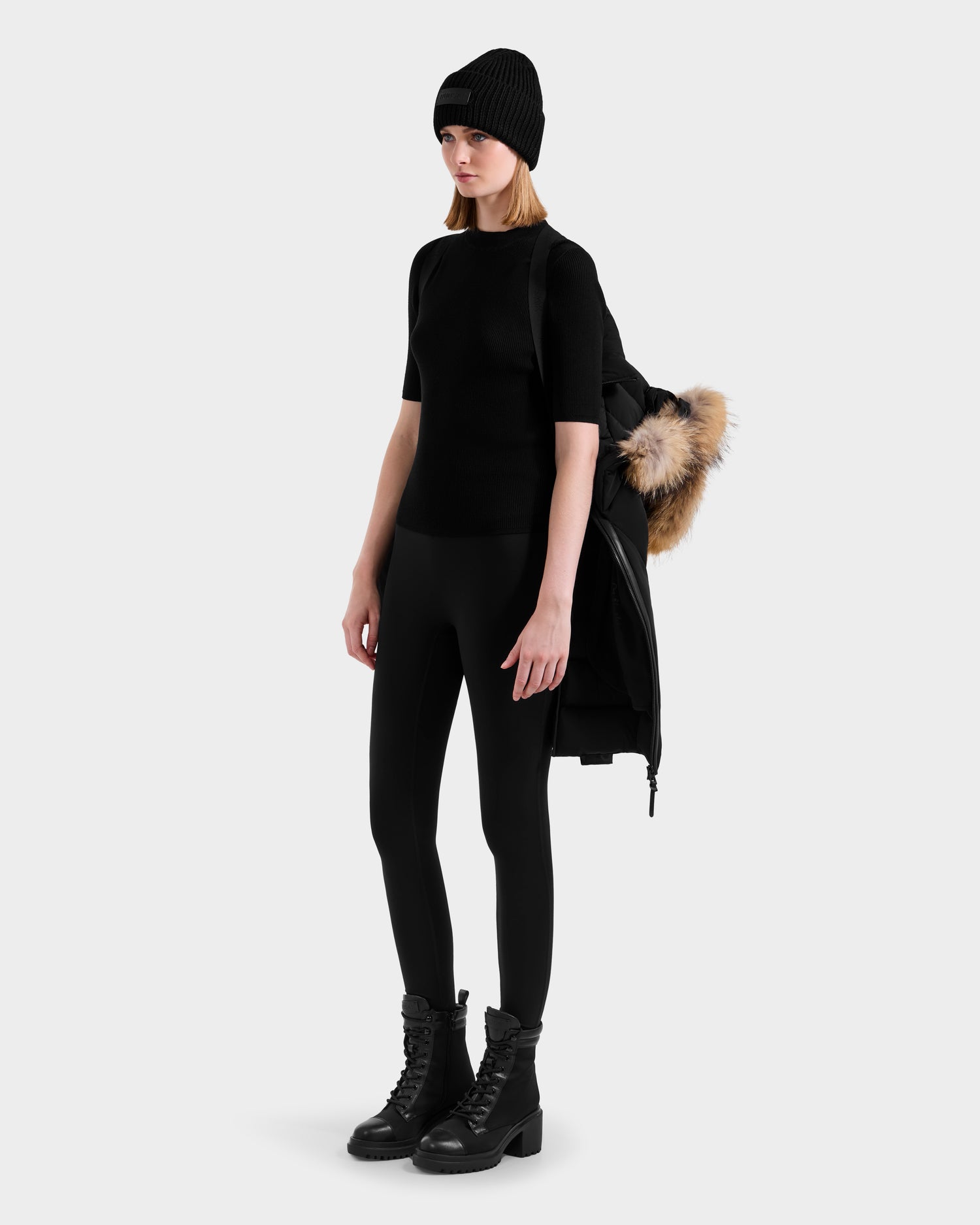 A woman stands against a plain background wearing the Rudsak MODA - 8125704 Black/Natural winter coat with fur trim draped over her shoulder, paired with a black beanie, short-sleeve top, leggings, and lace-up boots.