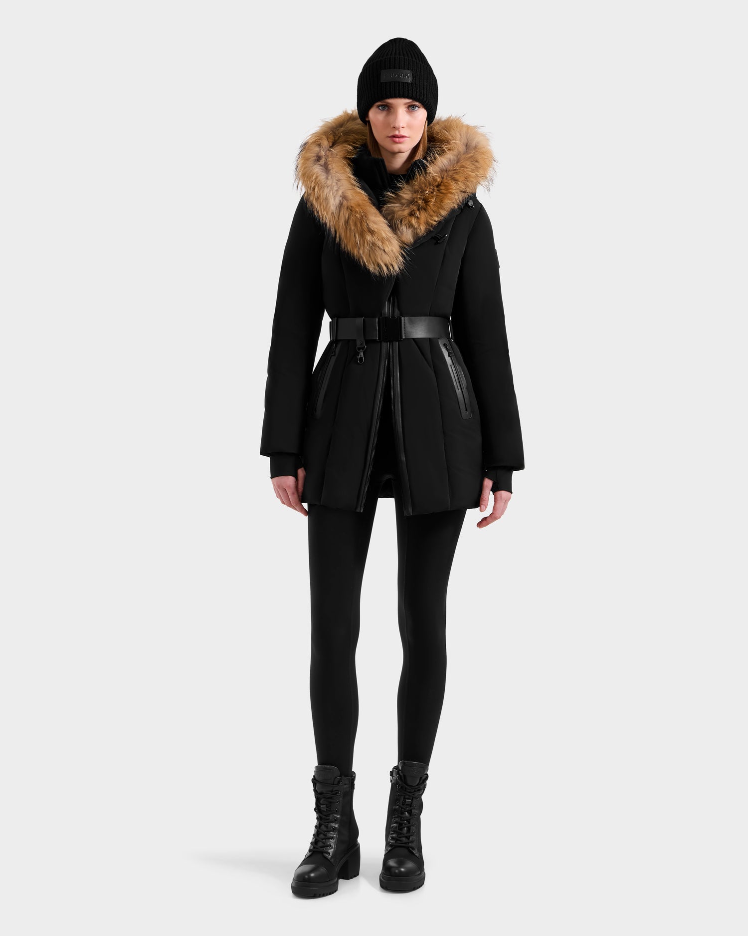 A woman faces forward wearing the Rudsak MODA - 8125704 Black/Natural winter coat with a large brown fur-lined hood, black leggings, lace-up boots, and a black knit beanie against a plain light background.