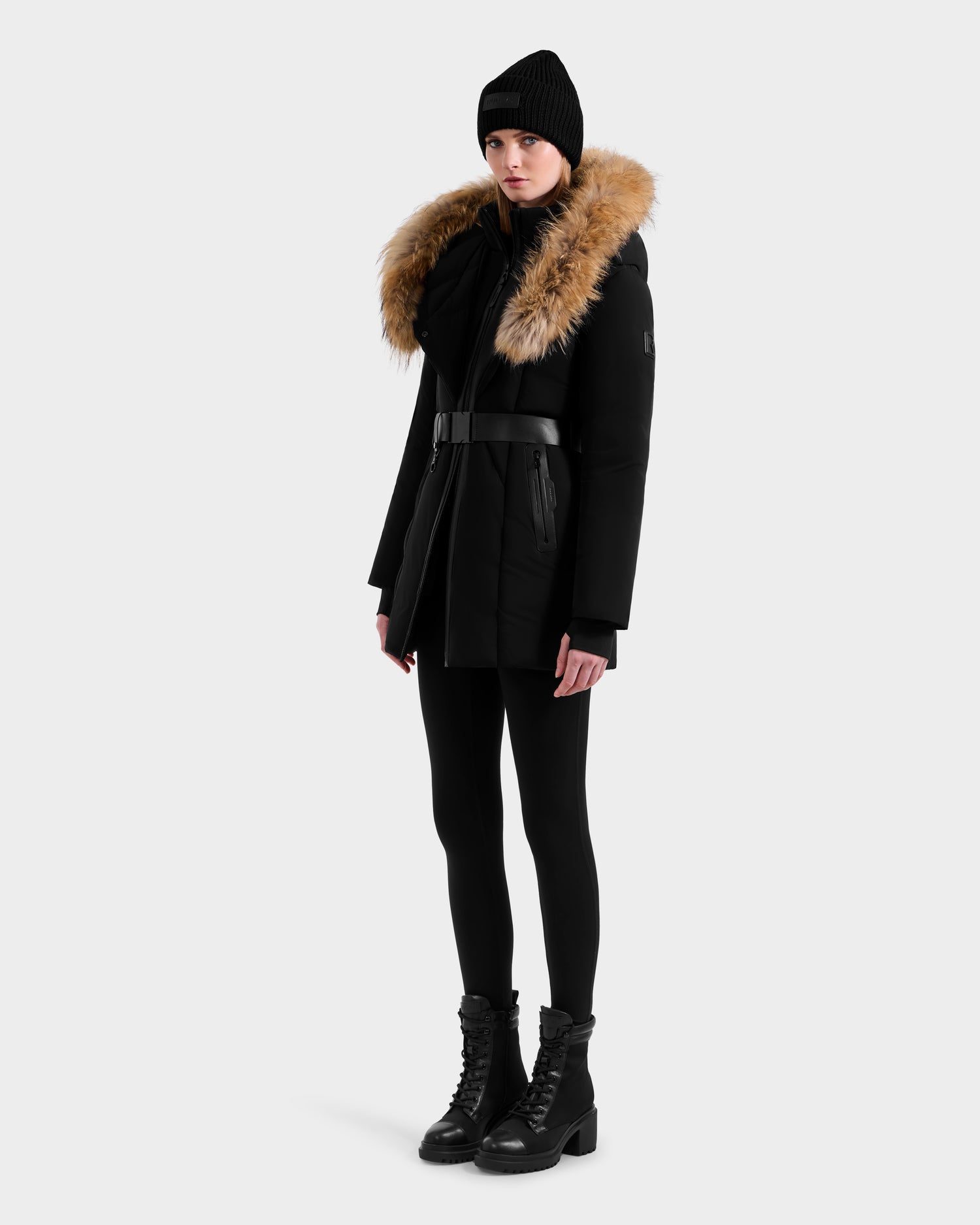 A woman wears the Rudsak MODA - 8125704 Black/Natural winter coat with a fur-trimmed hood, black beanie, leggings, and lace-up boots, standing against a plain white background.