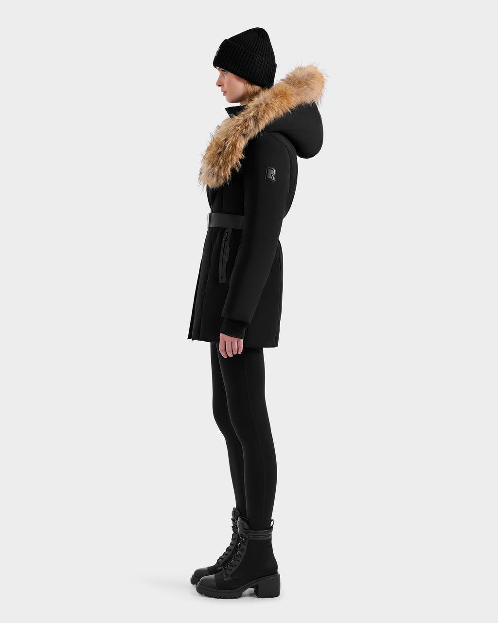 A person stands in profile against a light gray background wearing the Rudsak MODA - 8125704 Black/Natural down parka with tan fur-lined hood, paired with black leggings, lace-up boots, and a black knit beanie.