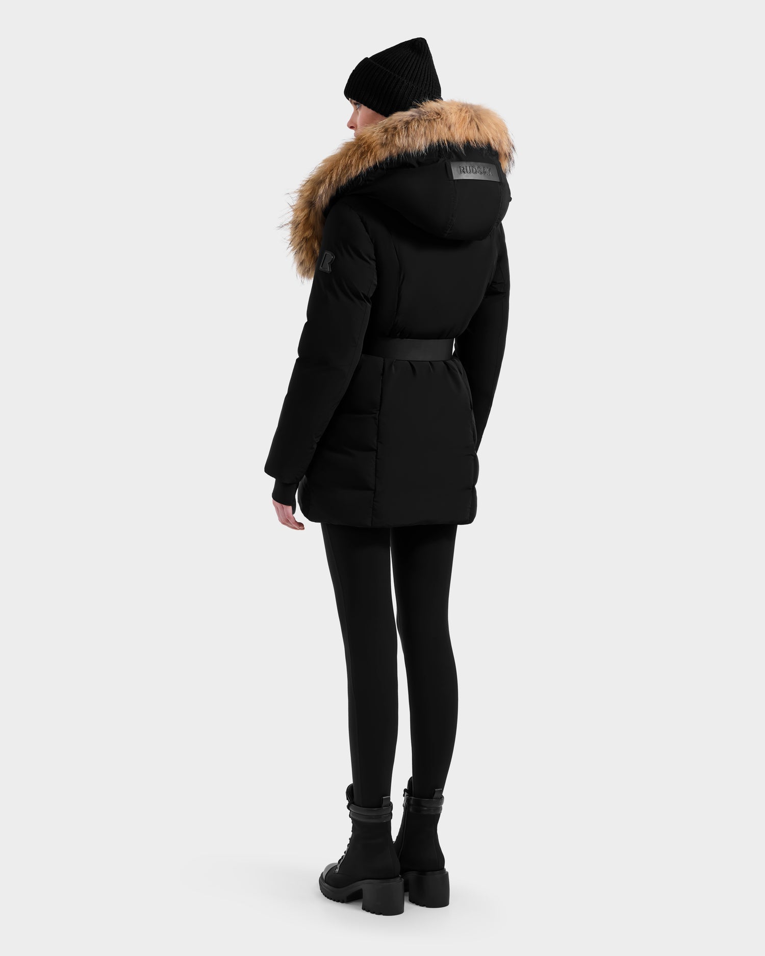 A person models the Rudsak MODA - 8125704 Black/Natural winter coat, featuring a fur-lined hood, against a plain light background, highlighting its timeless style and sustainable design.