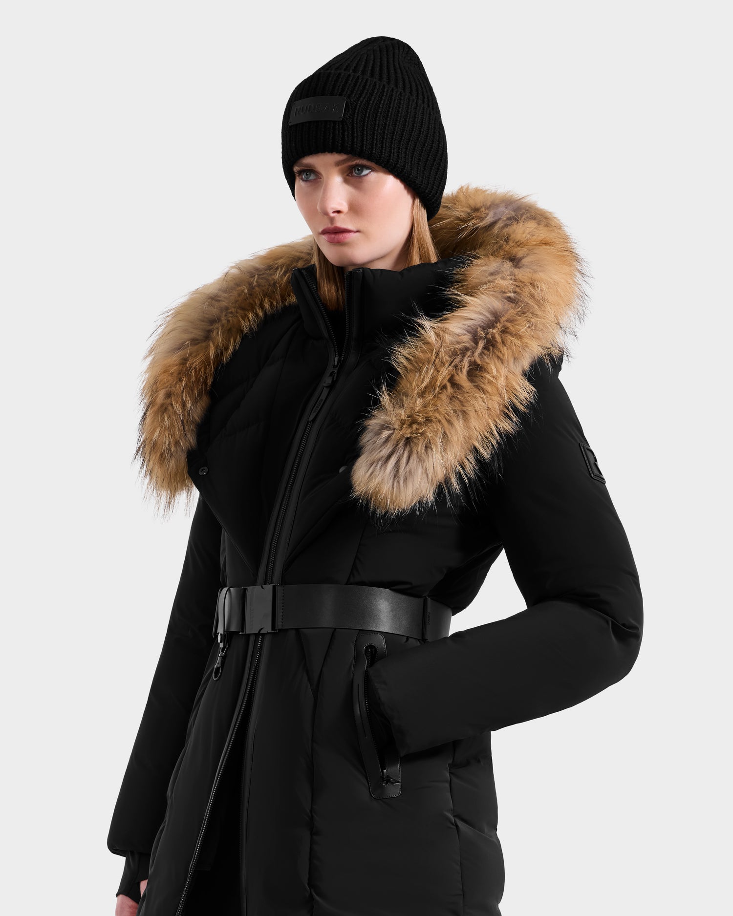 A woman wears the Rudsak MODA - 8125704 Black/Natural down parka with a fur-lined hood, paired with a black beanie and belt, standing against a light grey background and gazing seriously to the side.