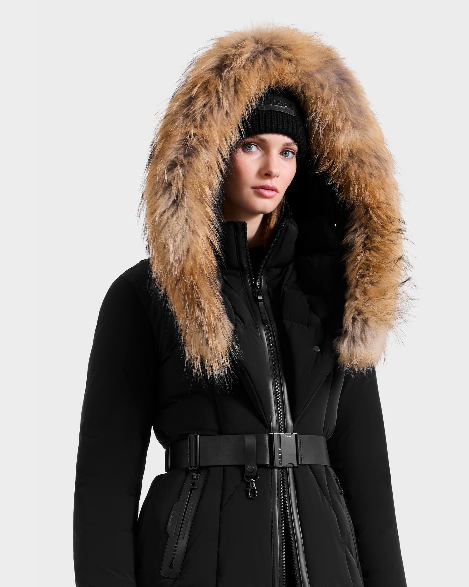 A person wearing Rudsak’s MODA - 8125704 Black/Natural coat with a wide, fur-trimmed hood and black knit beanie stands against a plain background, highlighting both warmth and sustainable design.