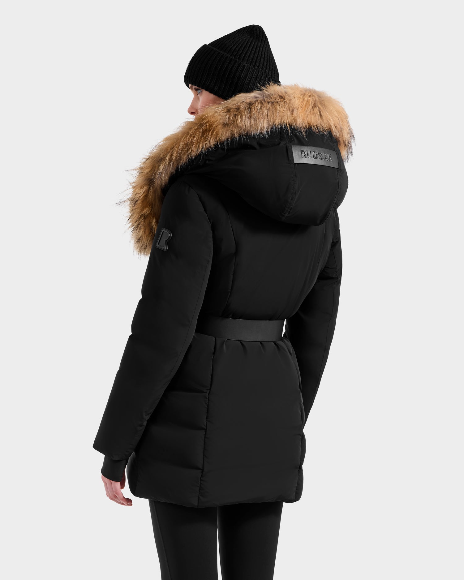 Shown from the back against a plain light background, a person is wearing the Rudsak MODA - 8125704 Black/Natural down parka with a fur-trimmed hood, paired with a black knit beanie and black pants.