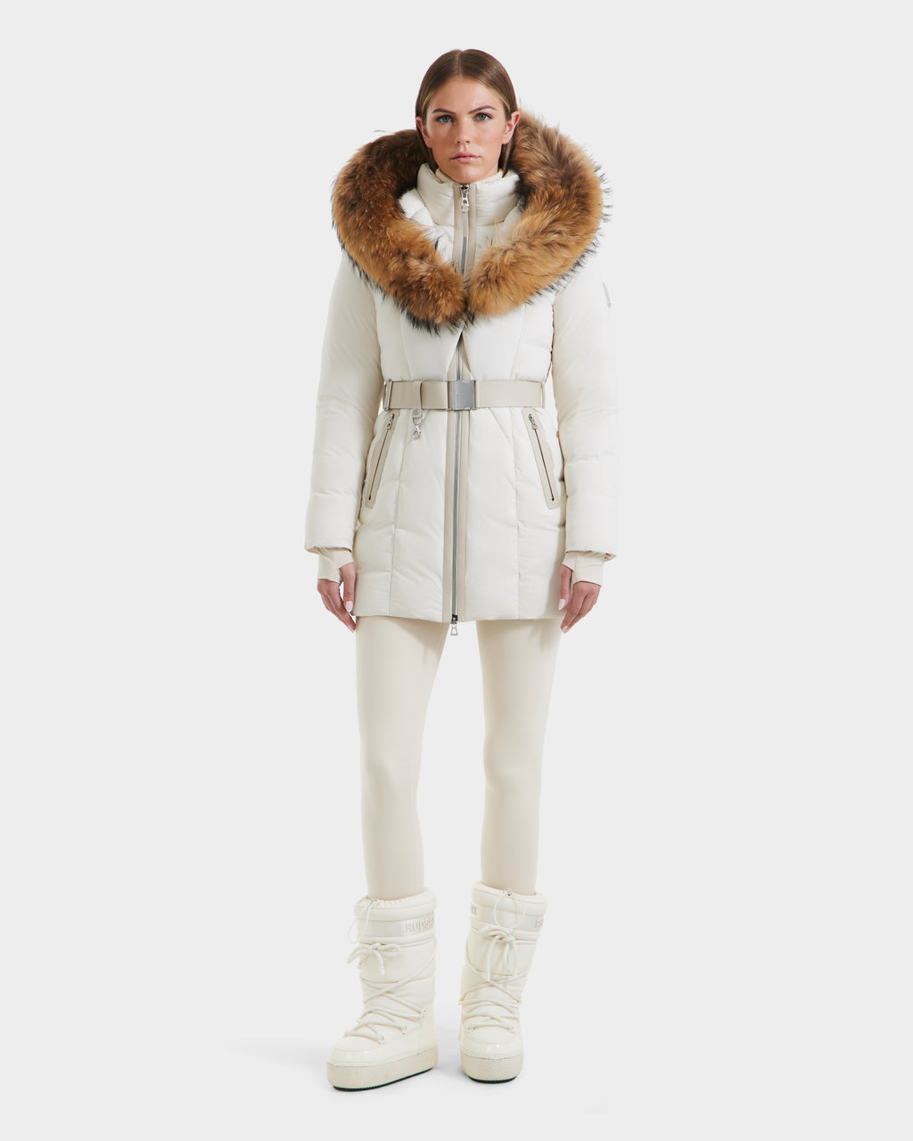 A woman faces forward in the Rudsak MODA - 8125704 Vanilla: a white belted winter coat with a large brown fur-trimmed hood, styled with white pants and tall snow boots—showcasing sustainable elegance against a plain background.