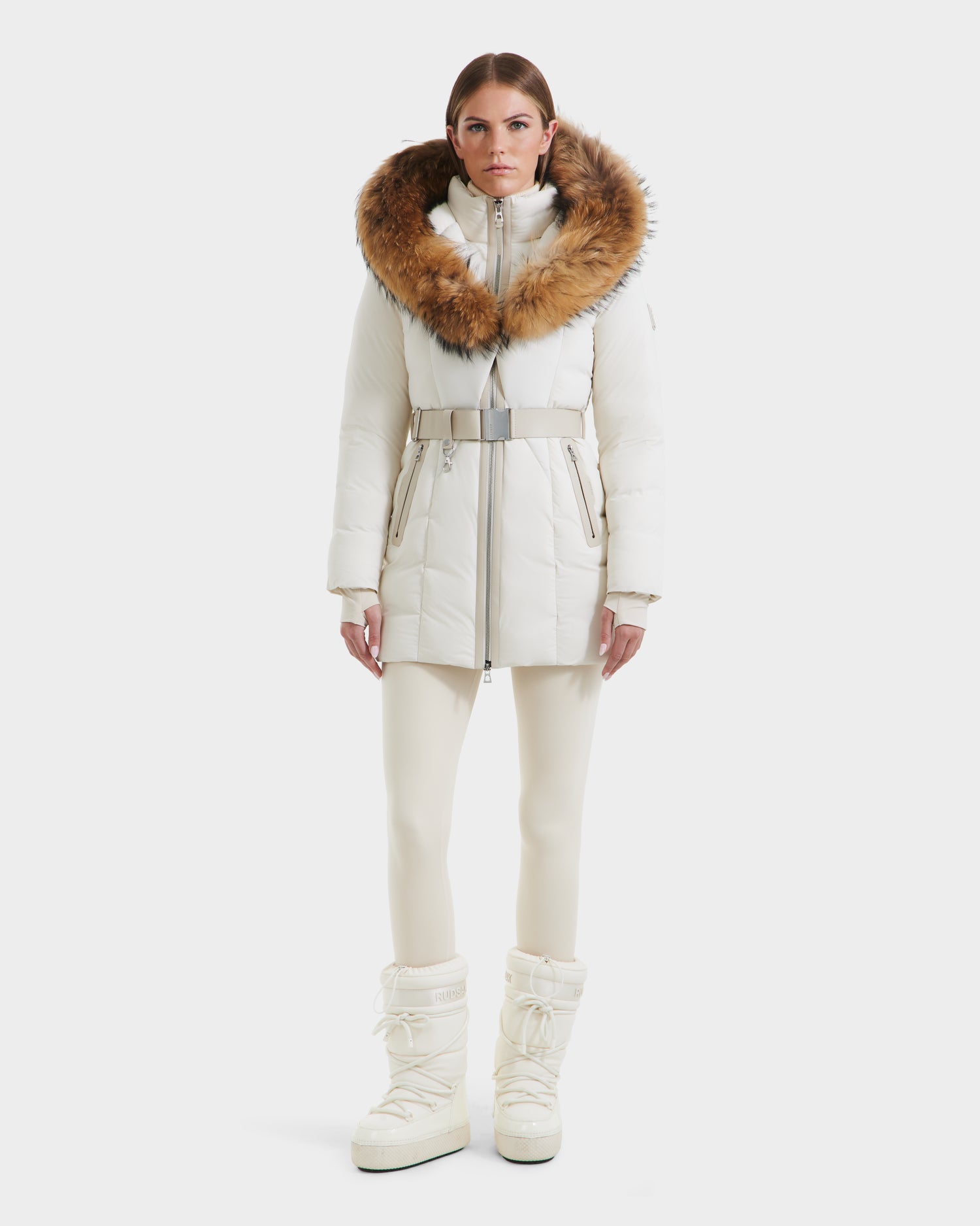 A woman faces forward in the Rudsak MODA - 8125704 Vanilla: a white belted winter coat with a large brown fur-trimmed hood, styled with white pants and tall snow boots—showcasing sustainable elegance against a plain background.