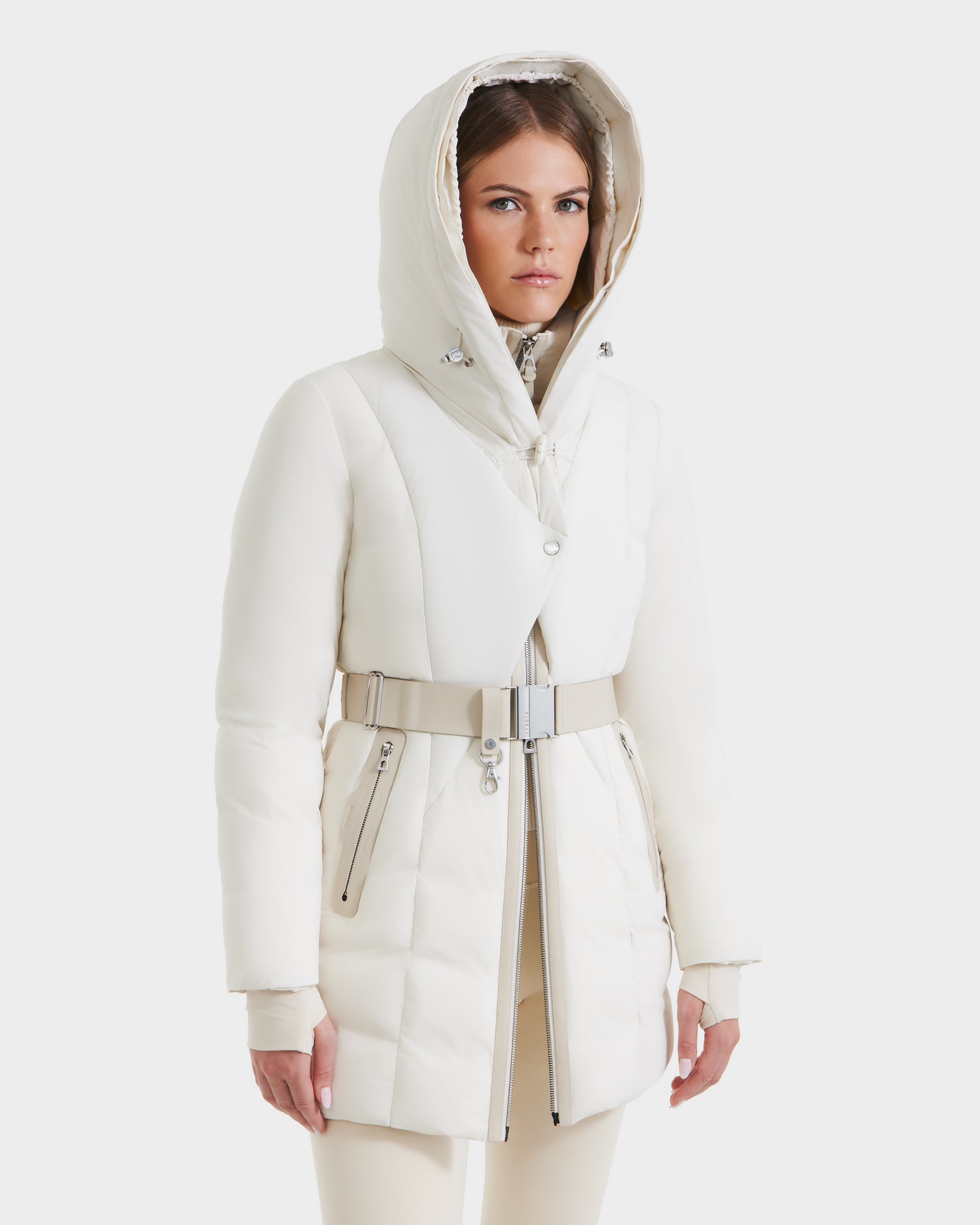 A woman in the Rudsak MODA - 8125704 Vanilla down parka with a belted waist and metallic zippers, paired with beige pants, stands against a white background—highlighting this modern, sustainable winter coat.