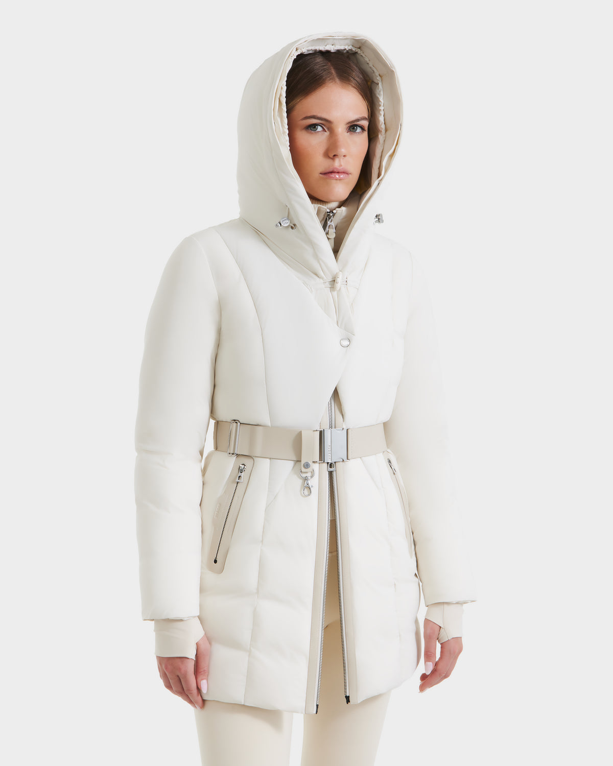 A woman in the Rudsak MODA - 8125704 Vanilla down parka with a belted waist and metallic zippers, paired with beige pants, stands against a white background—highlighting this modern, sustainable winter coat.