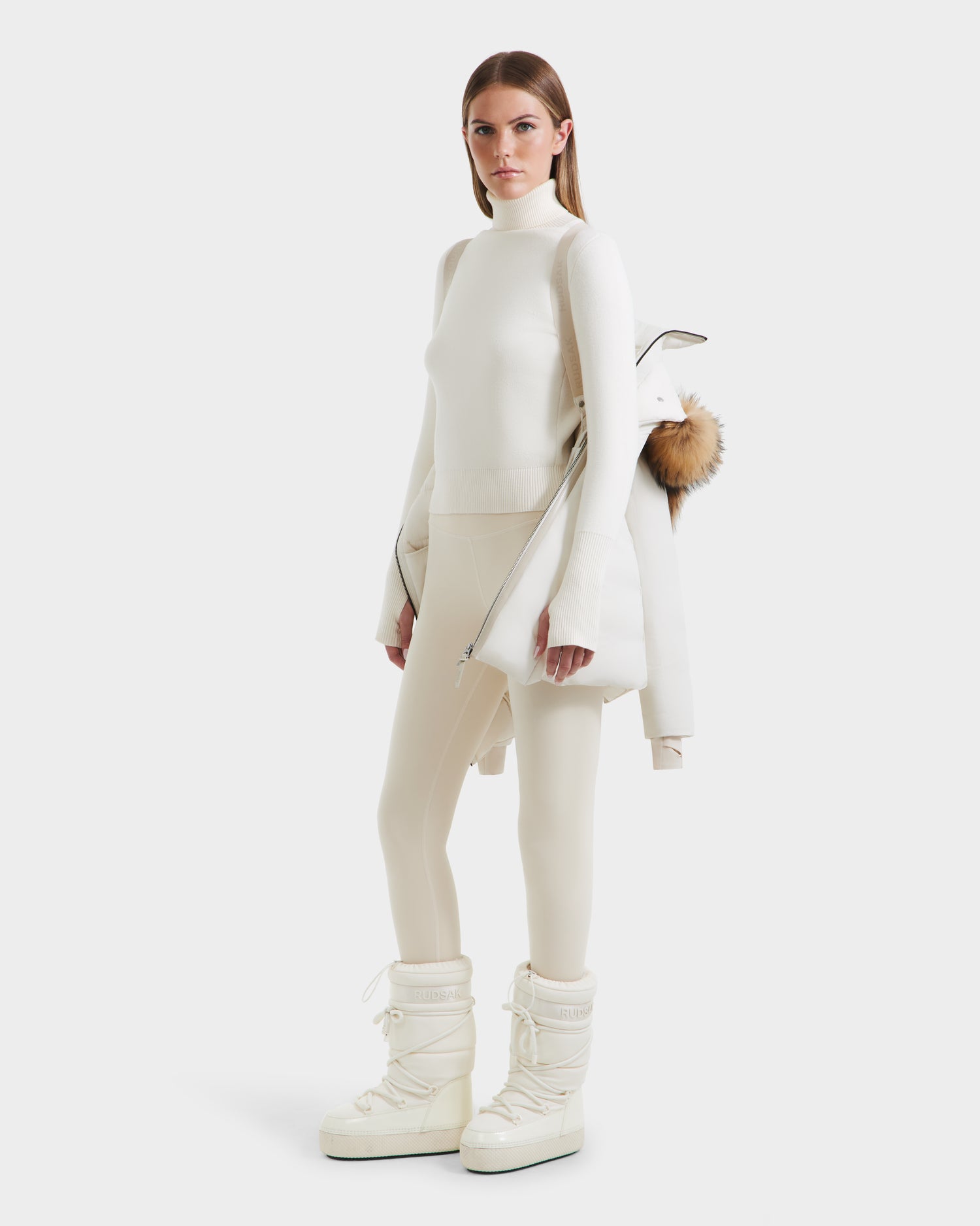 A woman stands against a white background in fitted cream attire and white winter boots, draping the Rudsak MODA - 8125704 Vanilla sustainable winter coat with fur-trimmed hood over her shoulders.
