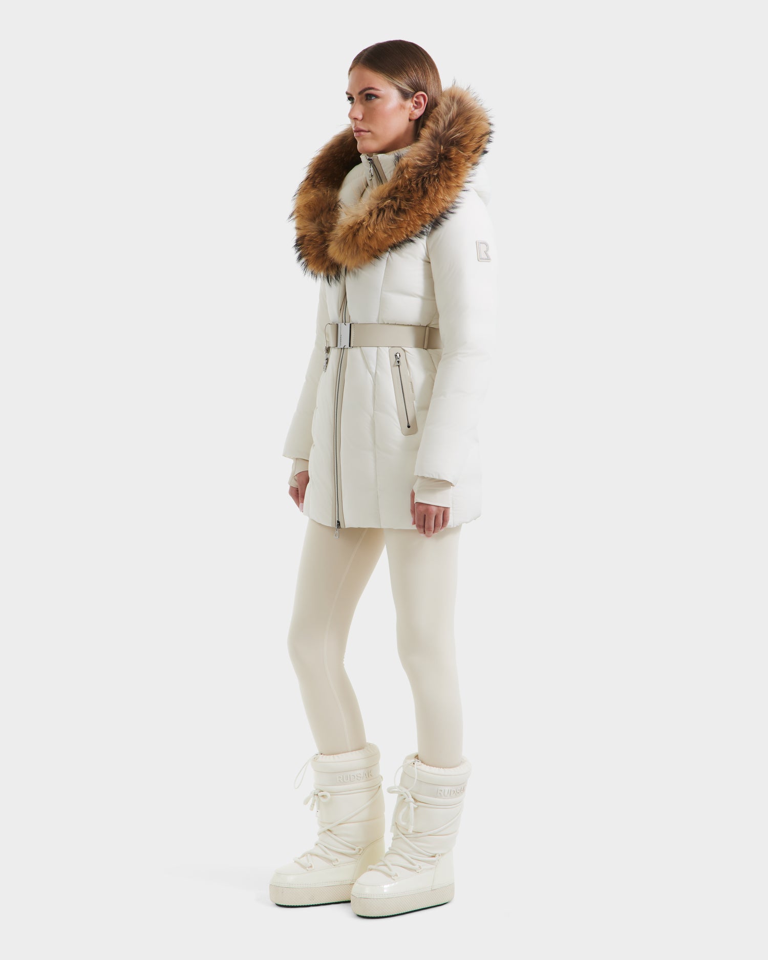 A woman in profile wears the Rudsak MODA - 8125704 Vanilla belted winter coat with a large brown fur collar, styled with white leggings and snow boots, set against a plain white background.