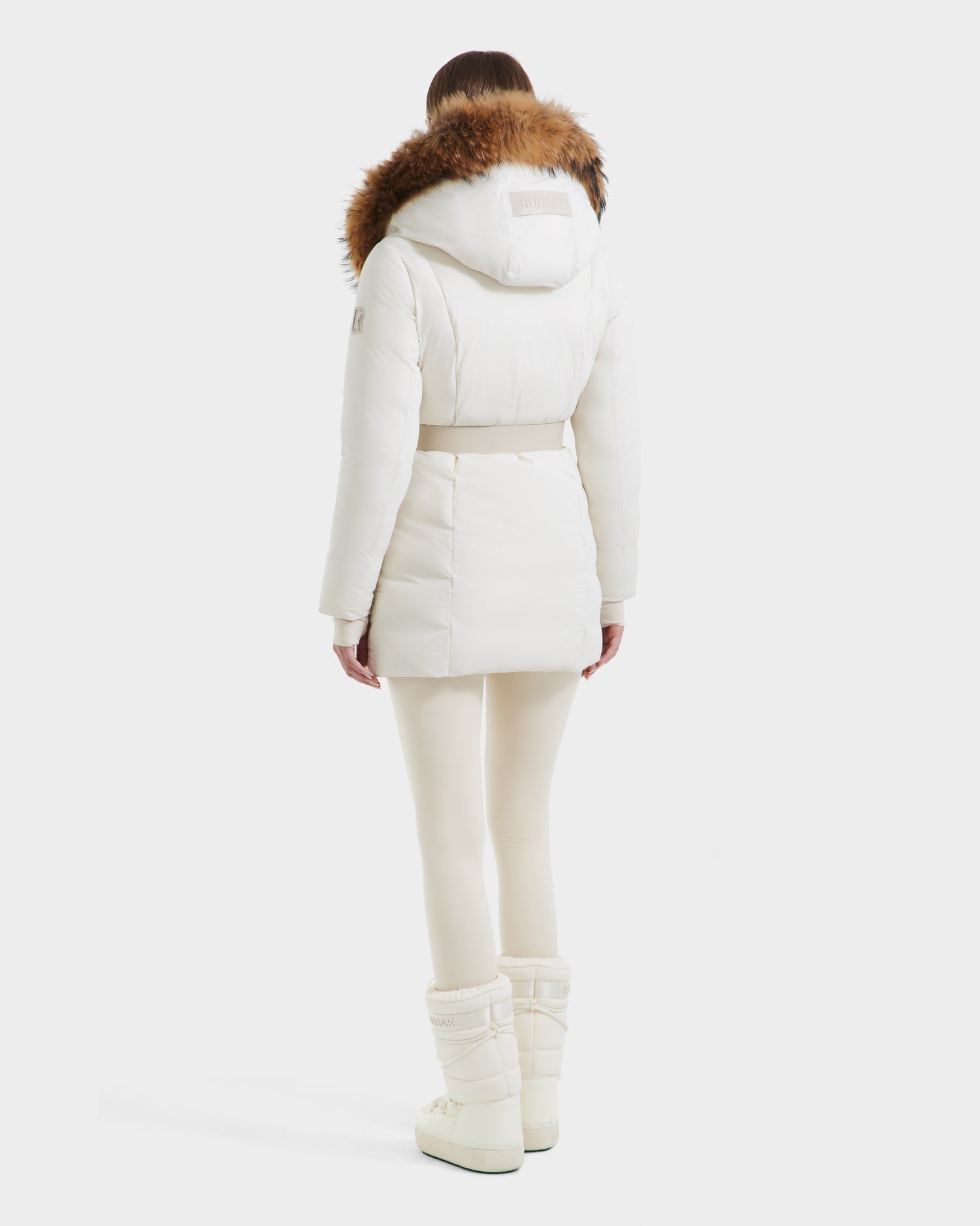 A person stands facing away, wearing the Rudsak MODA - 8125704 Vanilla white down parka with a brown fur-trimmed hood, white leggings, and boots against a plain backdrop—showcasing effortless sustainable style.