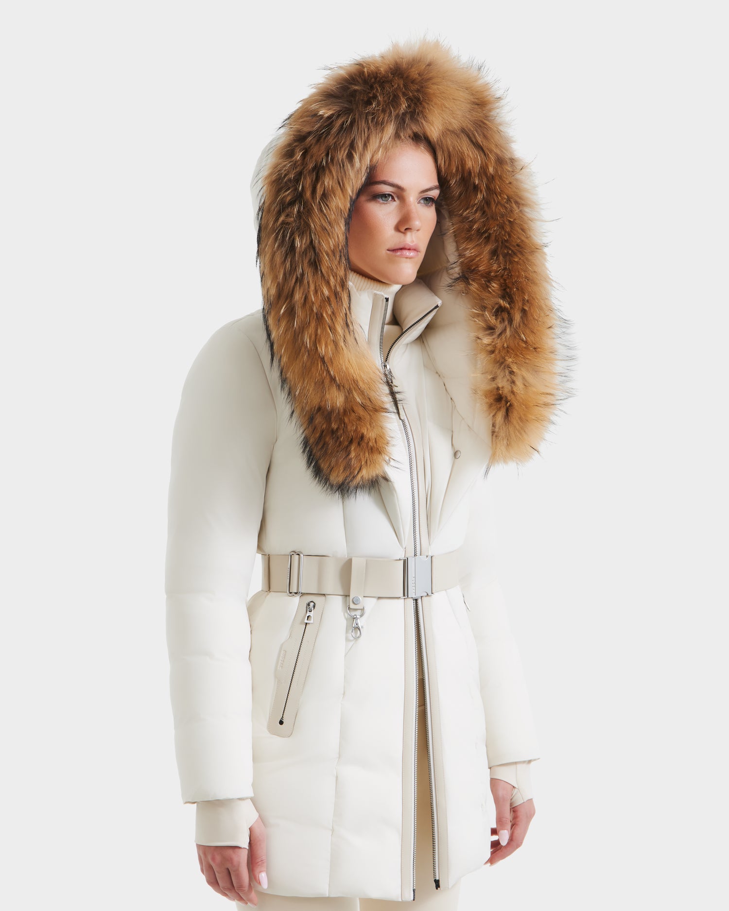 A woman models the Rudsak MODA - 8125704 Vanilla, a white belted winter coat with a large fur-trimmed hood. She stands against a plain white background with a neutral expression, highlighting its timeless and sustainable design.