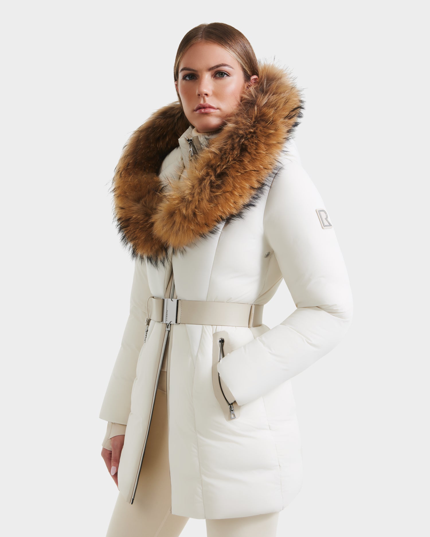 A woman wears the Rudsak MODA - 8125704 Vanilla winter coat, featuring a white belted design and a large brown fur-trimmed hood, standing confidently against a light gray background.