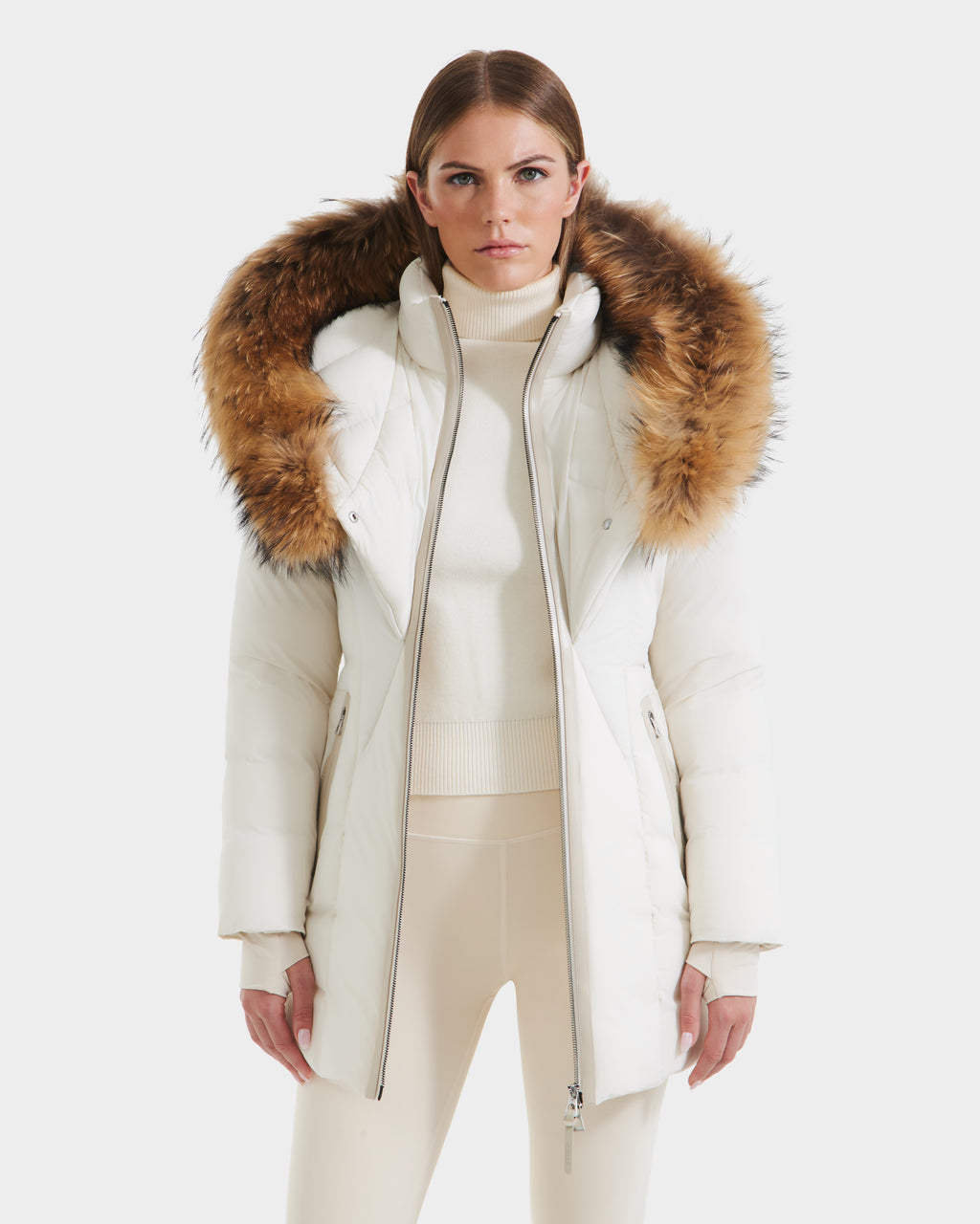 A woman wearing the Rudsak MODA - 8125704 Vanilla white down parka with a large brown fur-trimmed hood stands facing forward against a light background, paired with a cream turtleneck sweater and cream leggings.
