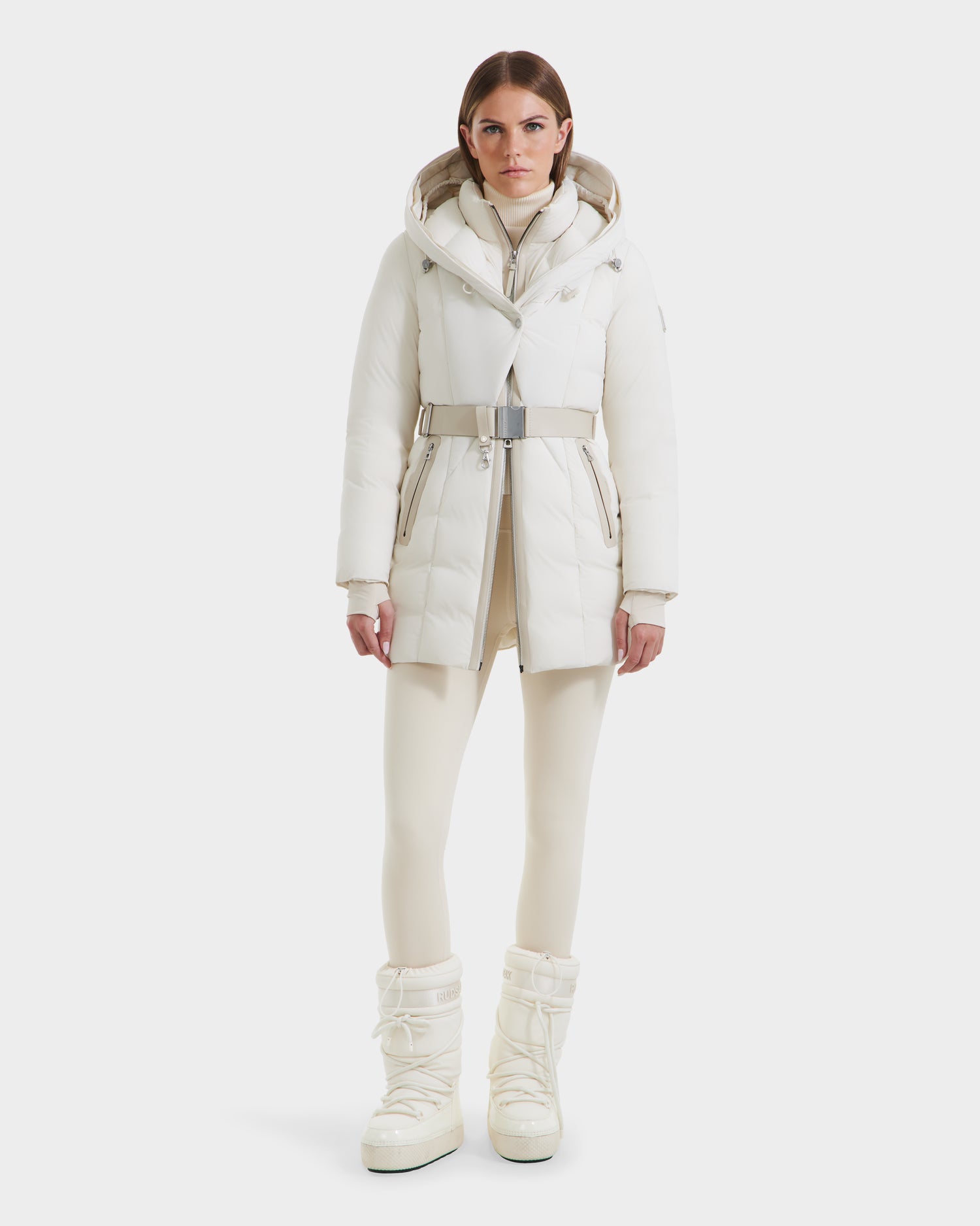 A woman with straight brown hair and a neutral expression wears the Rudsak MODA - 8125704 Vanilla white down parka with a belt, paired with matching leggings and tall winter boots, standing against a plain white background.