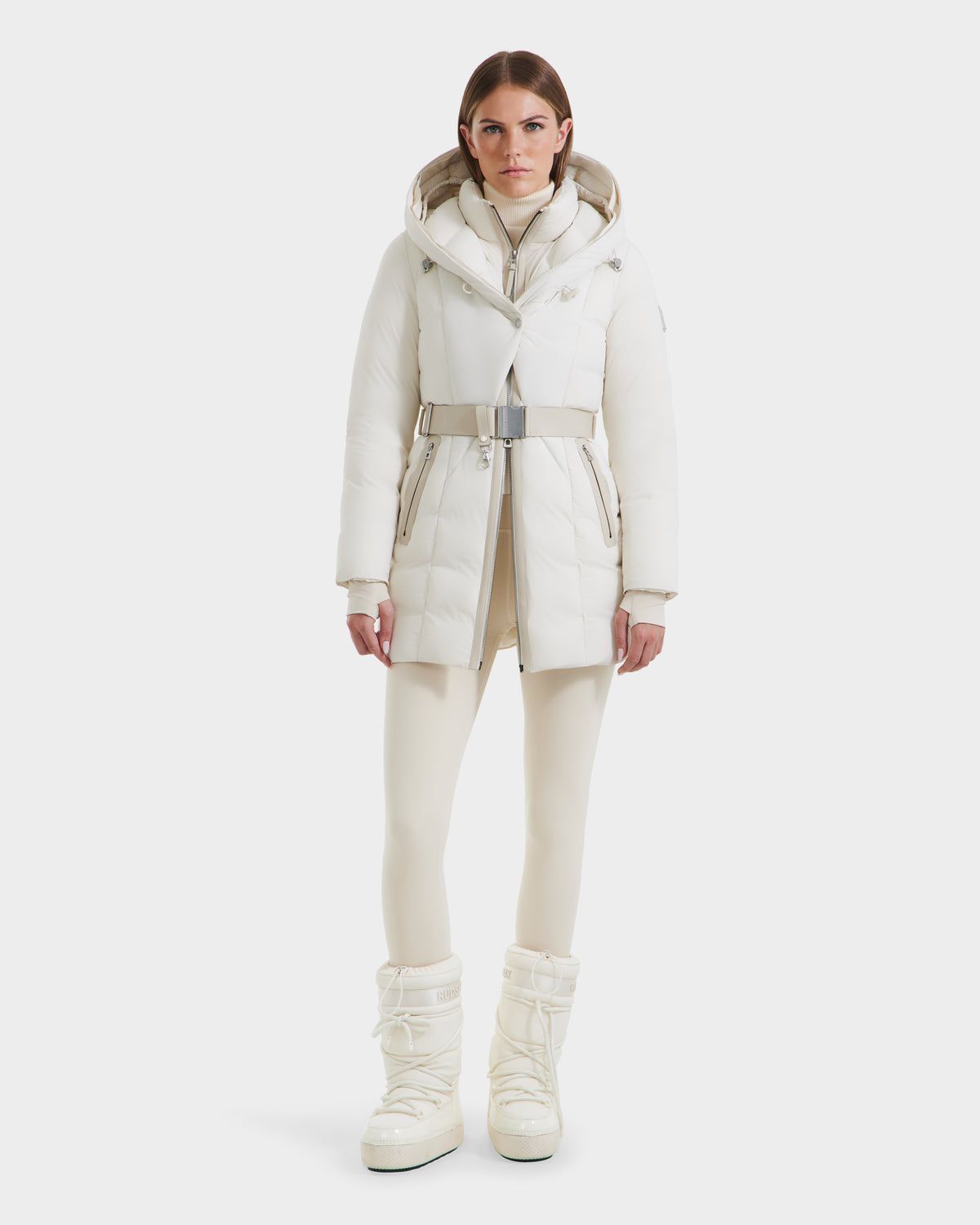A woman with straight brown hair and a neutral expression wears the Rudsak MODA - 8125704 Vanilla white down parka with a belt, paired with matching leggings and tall winter boots, standing against a plain white background.