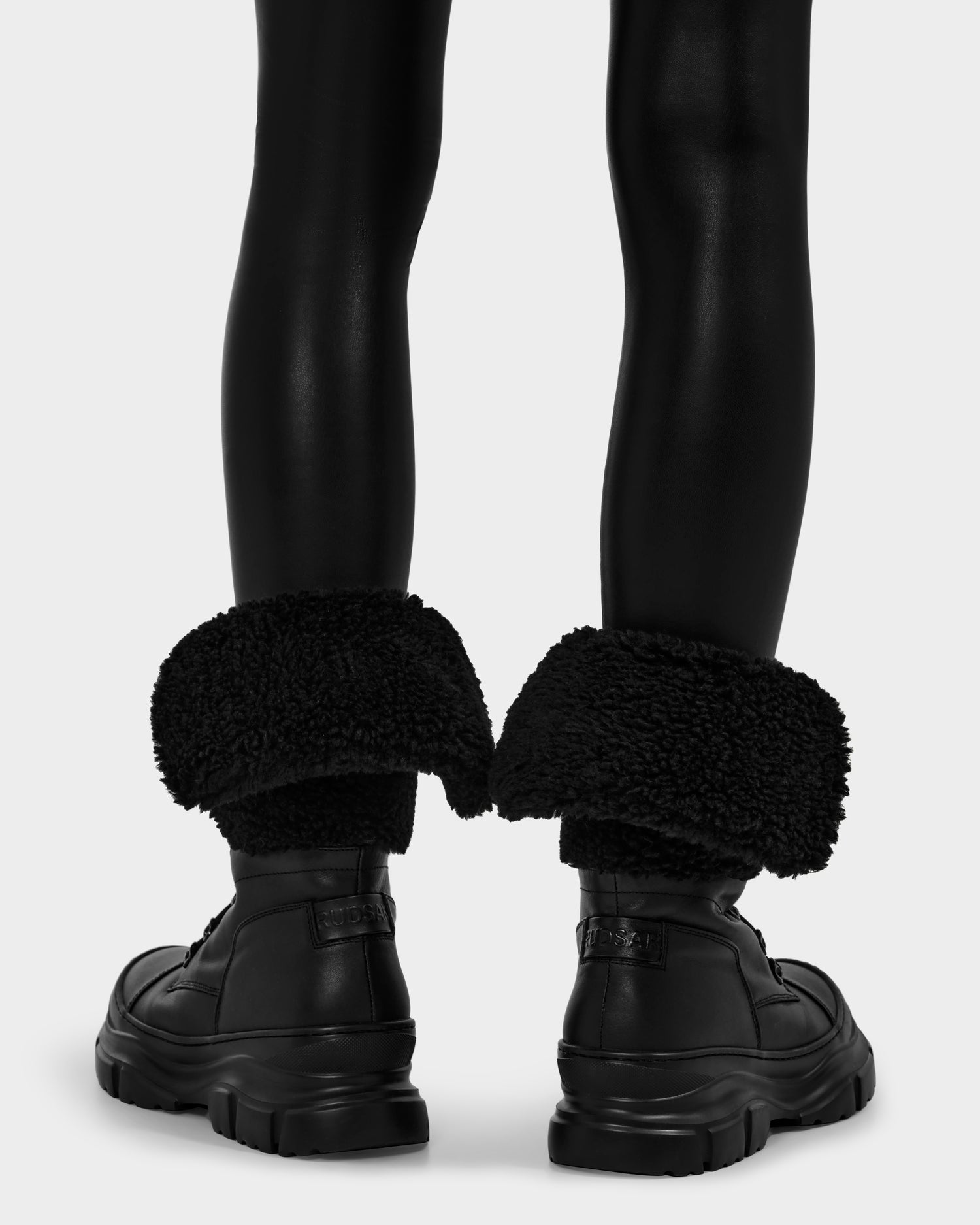 Someone wearing Rudsak MODENA - 8225024 Black chunky-soled winter boots with thick faux shearling cuffs and shiny black leggings, shown from the back against a plain light background.
