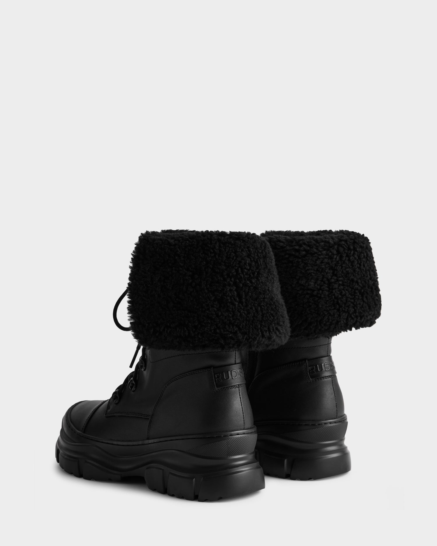 The MODENA - 8225024 Black winter boots by Rudsak feature chunky soles and folded-down faux shearling cuffs, crafted in black leather. Shown from the back and angled to the side against a plain light background.