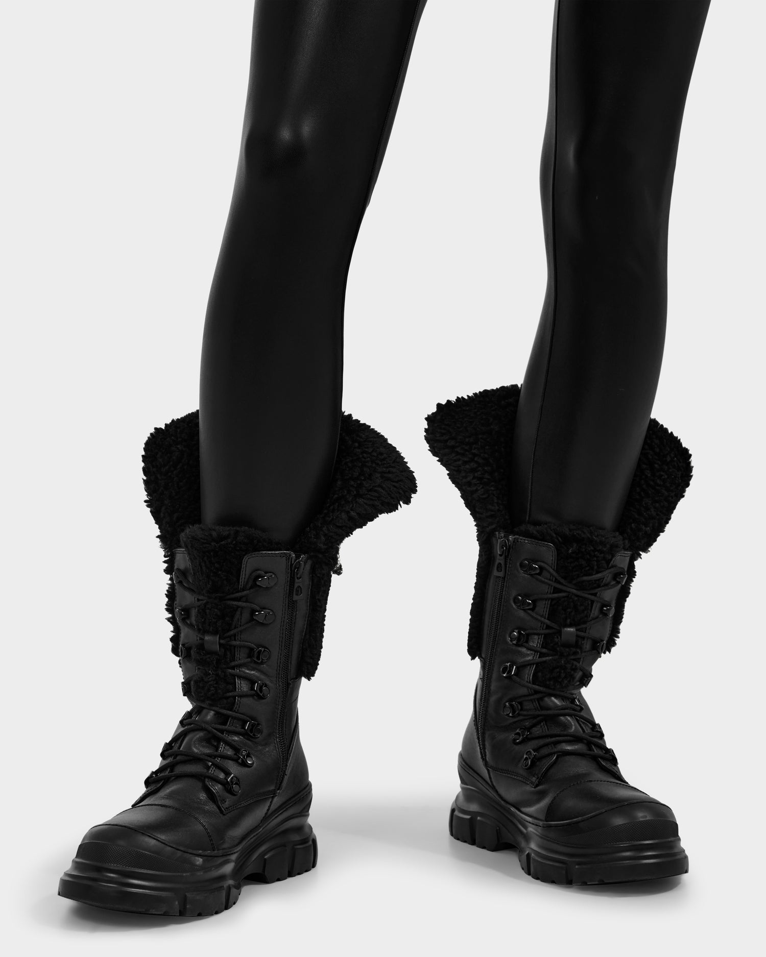 A person wears Rudsak MODENA - 8225024 Black boots with thick soles and fluffy faux shearling collars, paired with shiny black leggings, standing against a light background.