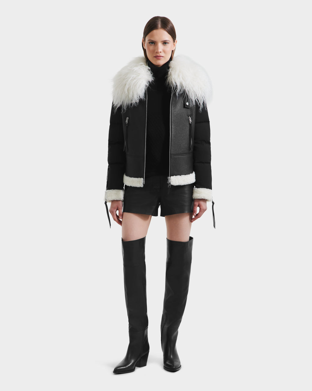 A woman faces forward against a plain white background, wearing the Rudsak MONKA - 8125629 Black women’s shearling jacket with a large Mongolian collar, a black turtleneck, black leather shorts, and knee-high boots.