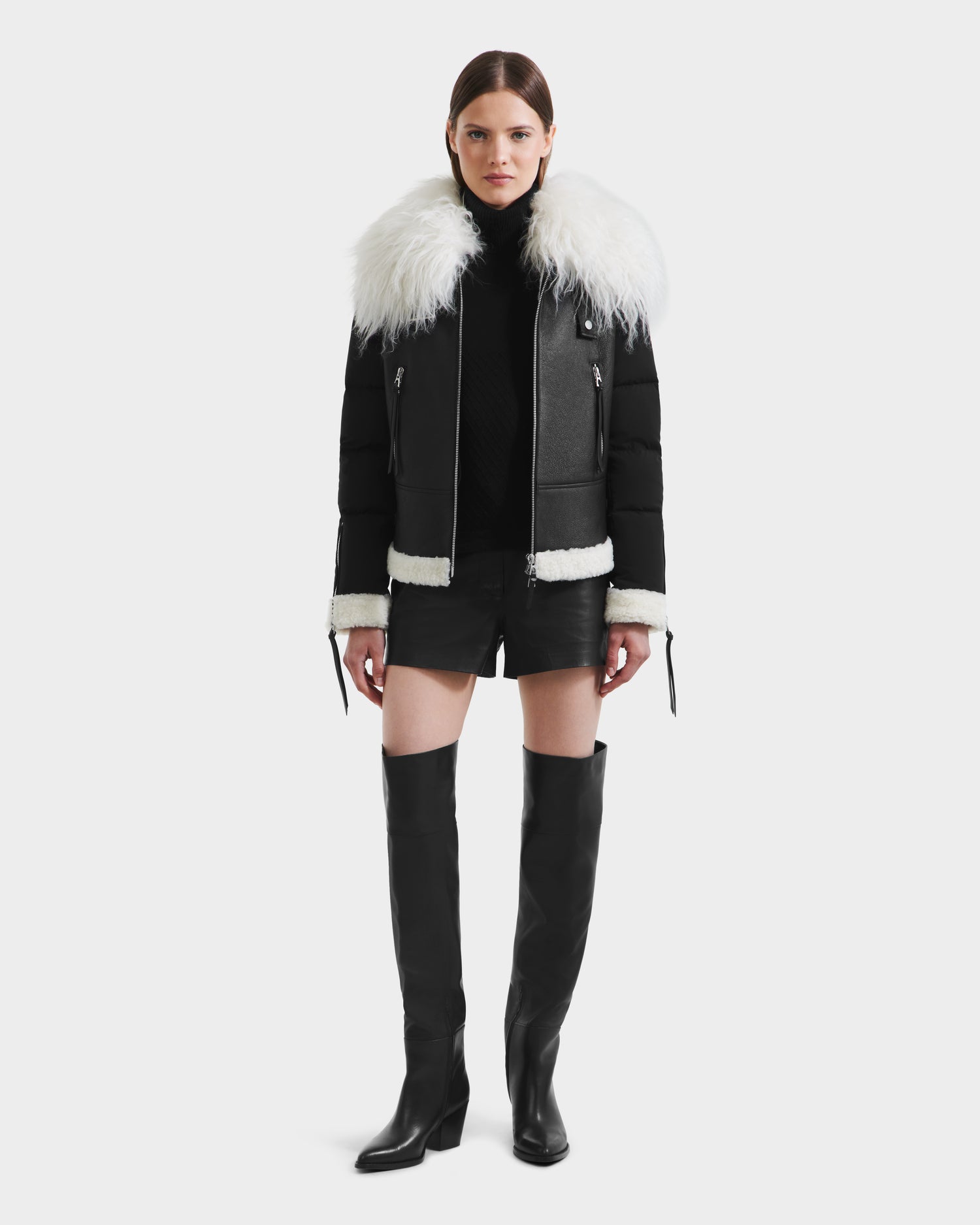 A woman faces forward against a plain white background, wearing the Rudsak MONKA - 8125629 Black women’s shearling jacket with a large Mongolian collar, a black turtleneck, black leather shorts, and knee-high boots.