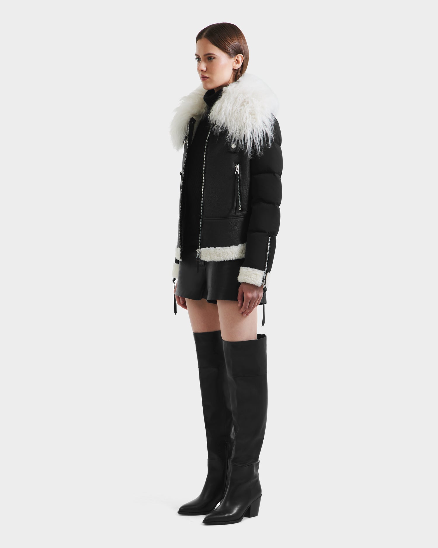 A woman wears the Rudsak MONKA - 8125629 Black women’s shearling jacket with a large Mongolian shearling collar, styled with black shorts and knee-high boots, standing against a plain white background.