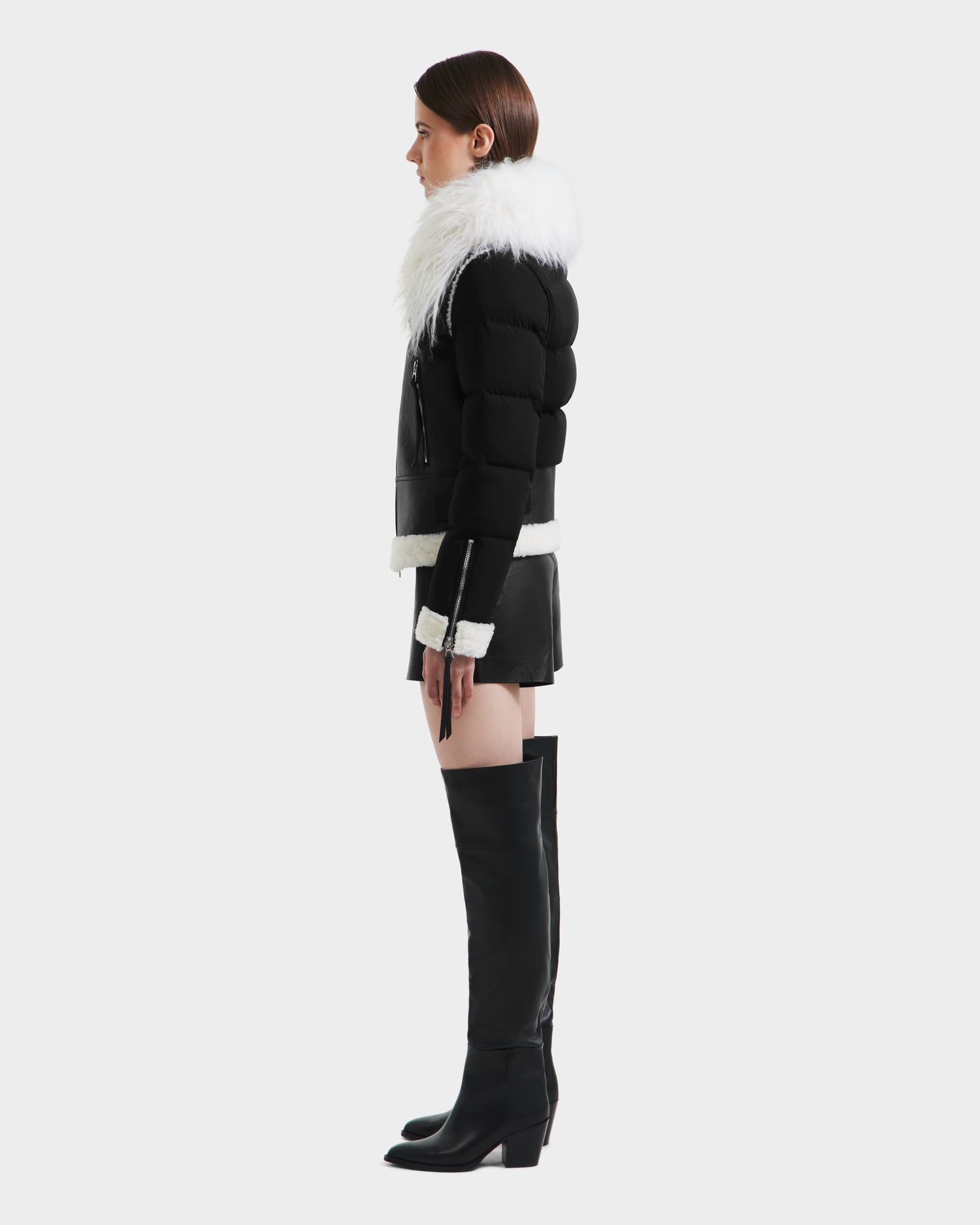 A woman in profile models the Rudsak MONKA - 8125629 Black puffer jacket with a large Mongolian shearling collar, styled with a black mini skirt and knee-high boots, highlighting its lightweight warmth against a plain light background.