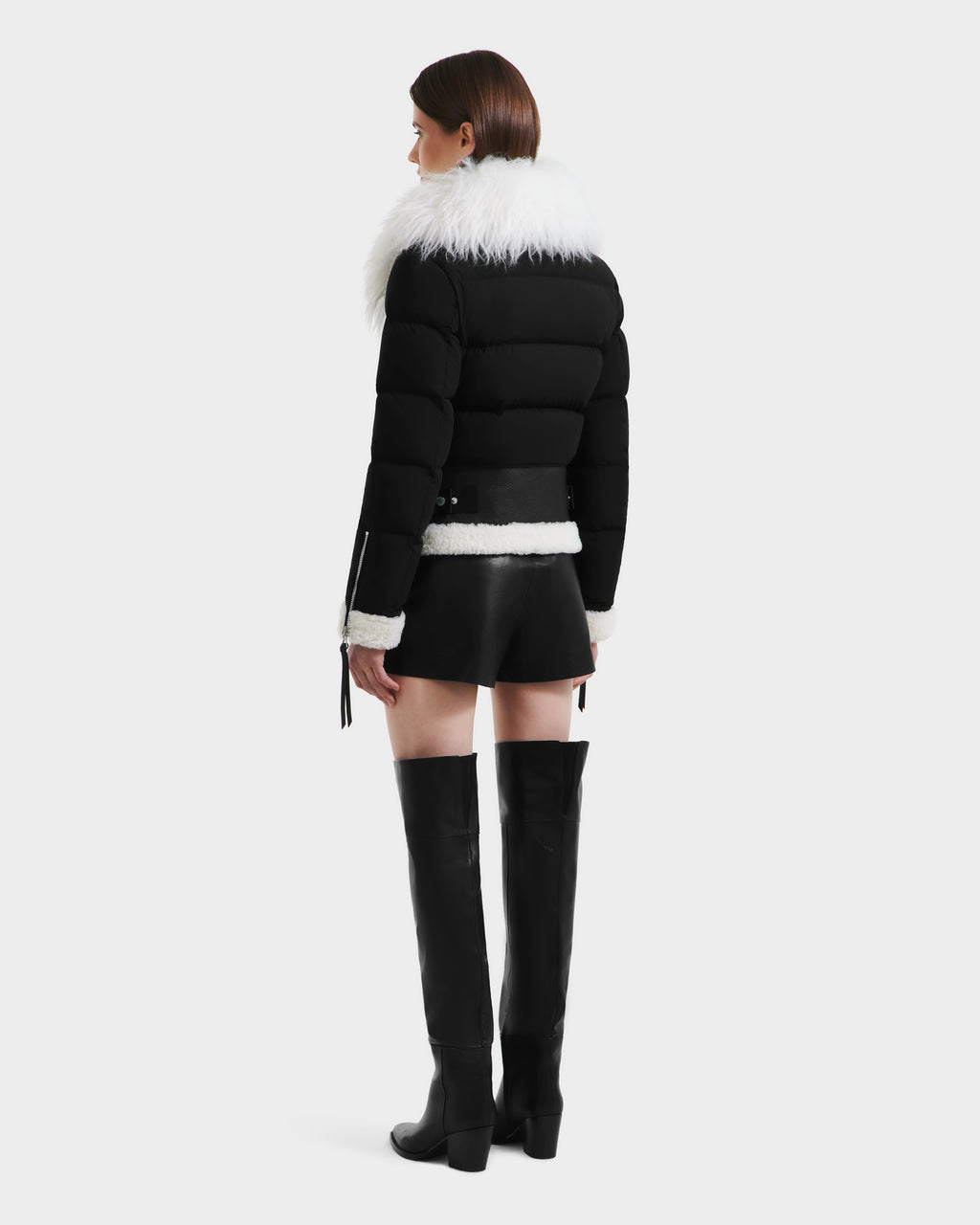 A woman faces away, wearing Rudsak's MONKA - 8125629 Black puffer jacket with a large Mongolian shearling collar, styled with black shorts and knee-high boots. The bold look stands out against a plain white background.
