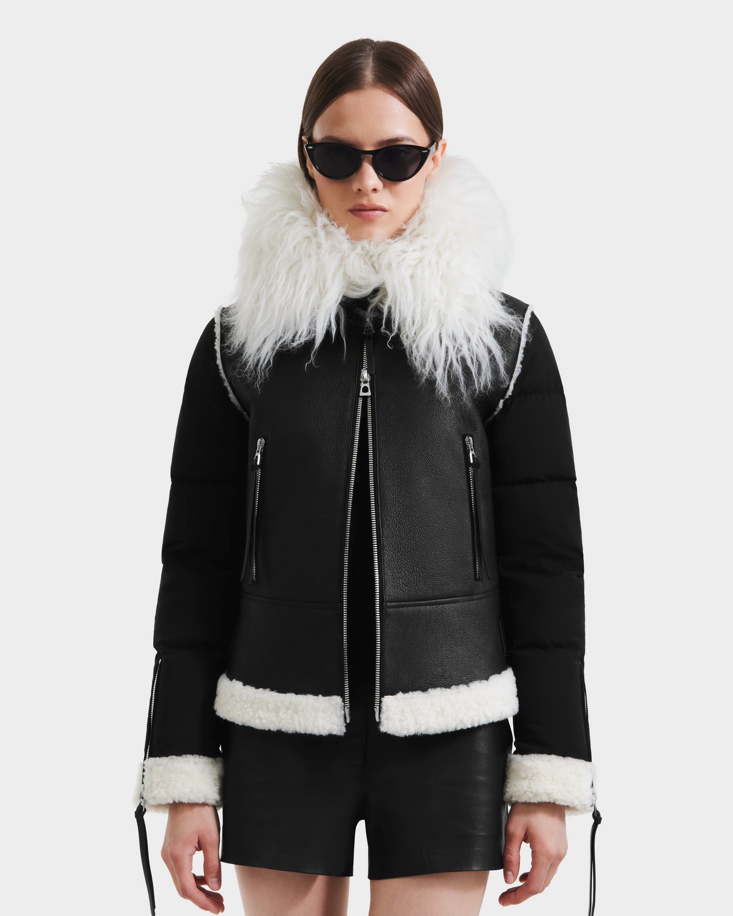 A woman wearing Rudsak’s MONKA - 8125629 Black women’s shearling jacket with a Mongolian shearling collar and black sunglasses stands against a white background, pairing the look with black shorts and a neutral expression.
