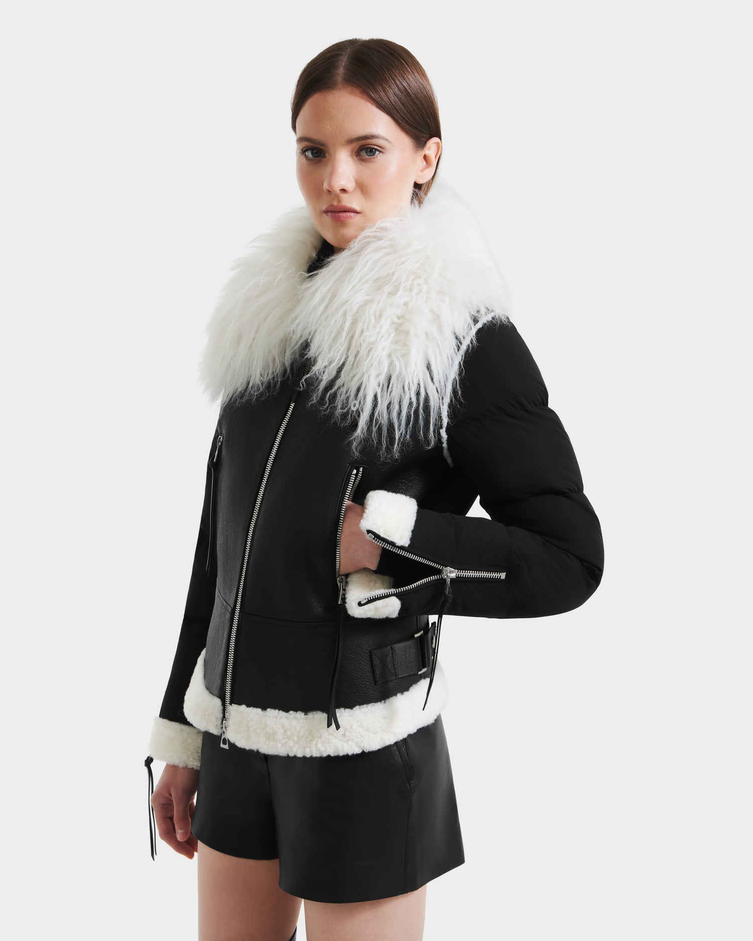 A woman models the Rudsak MONKA - 8125629 Black women’s shearling jacket with a bold Mongolian shearling collar and white trim, pairing it with black shorts against a plain background to highlight its lightweight warmth and stylish look.