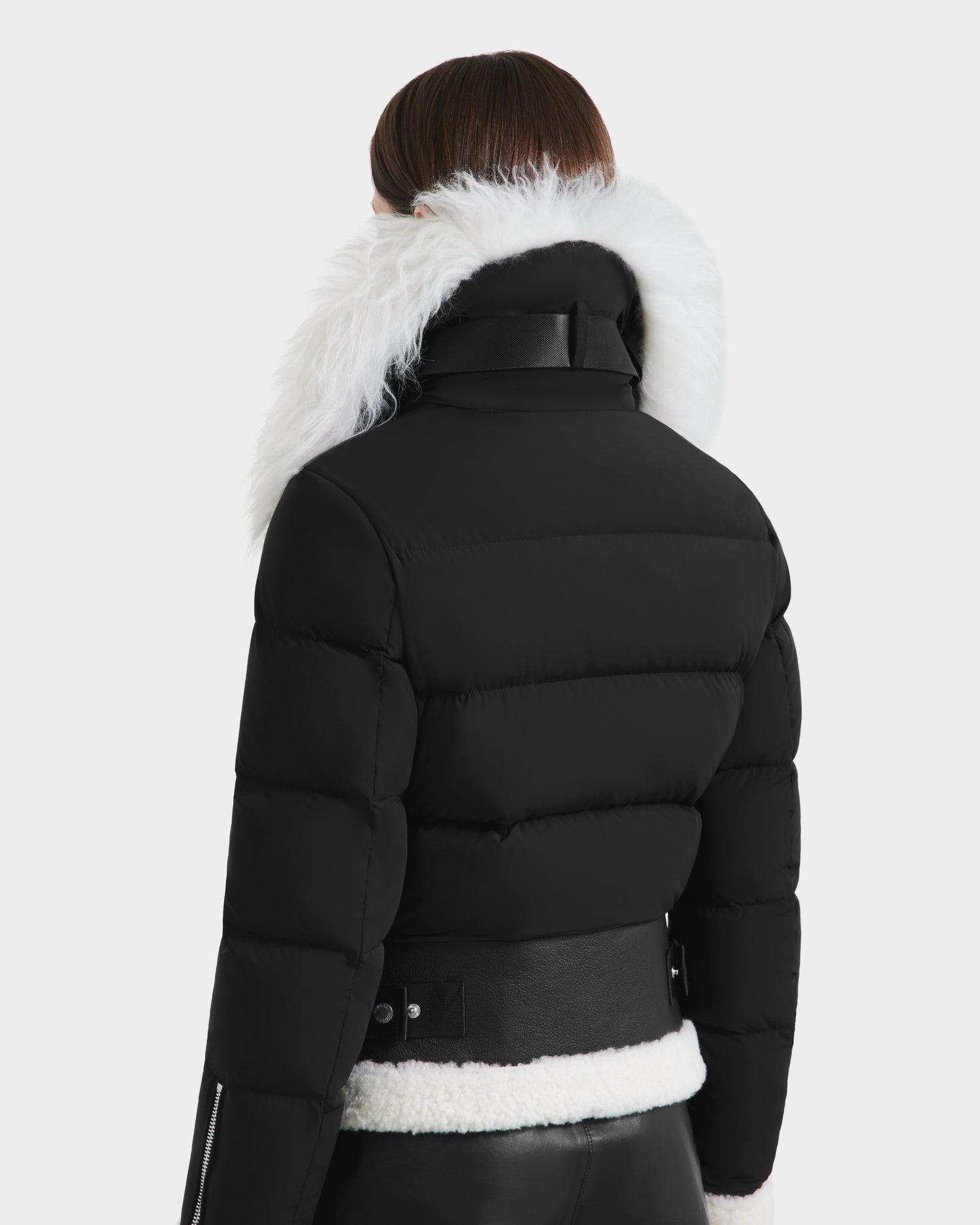 A person with straight brown hair wears the Rudsak MONKA - 8125629 Black women’s shearling jacket, featuring a Mongolian shearling collar, leather buckle waist details, and lightweight warmth with a high white faux fur-trimmed collar.