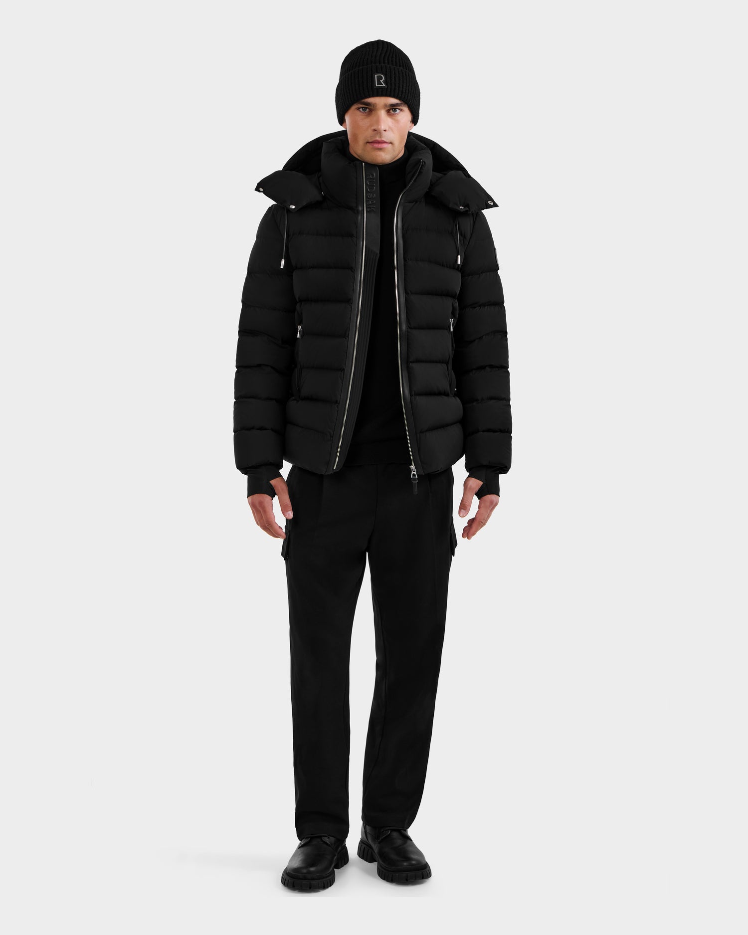 A person stands facing forward, wearing the Rudsak NAVAN - 6125598 Black men's quilted down puffer with RDS-certified fill, paired with a black beanie, pants, and shoes against a plain light background.
