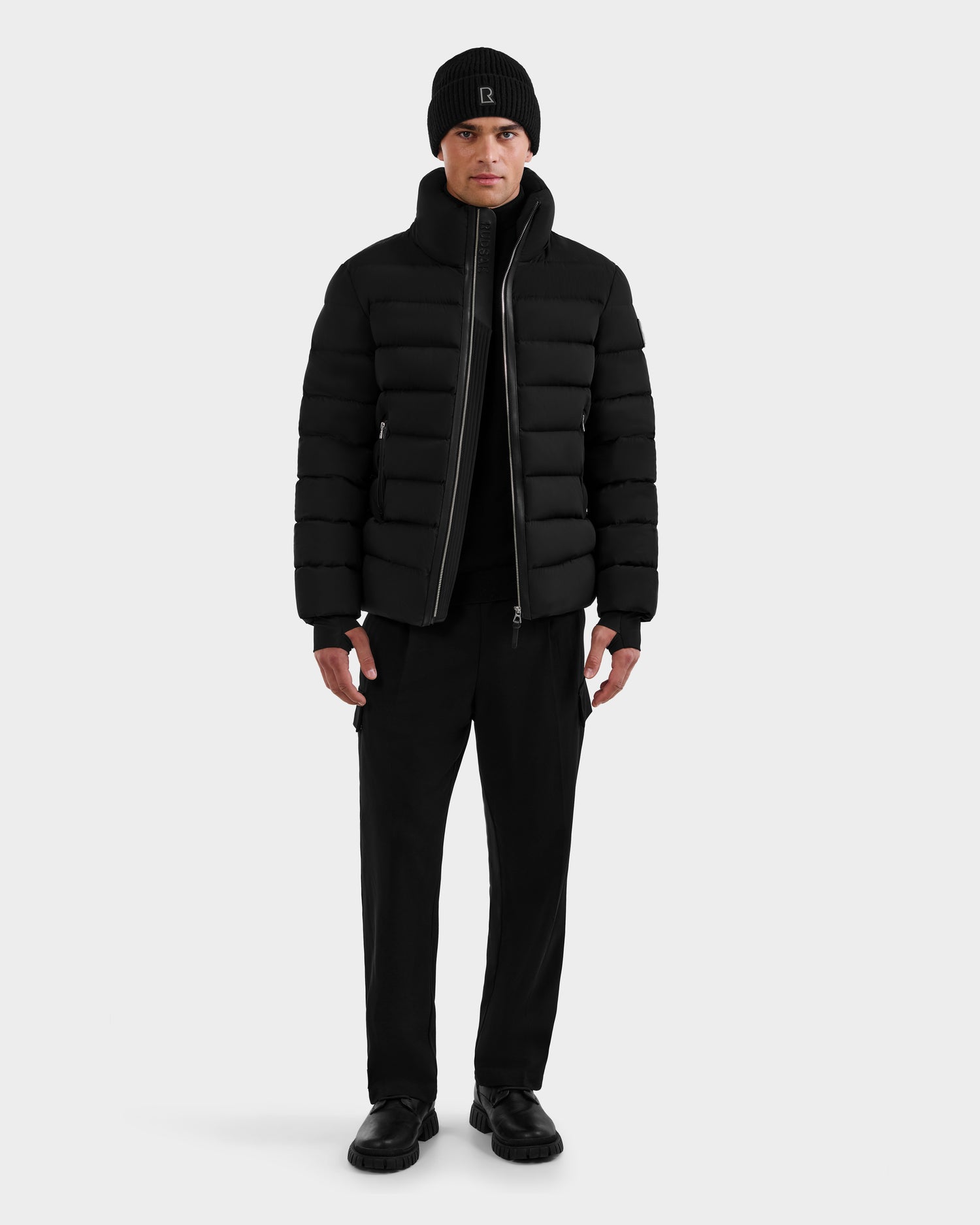 A person faces forward in a NAVAN - 6125598 Black men's quilted down puffer jacket by Rudsak, paired with a black beanie, sweater, pants, and shoes against a plain white background.