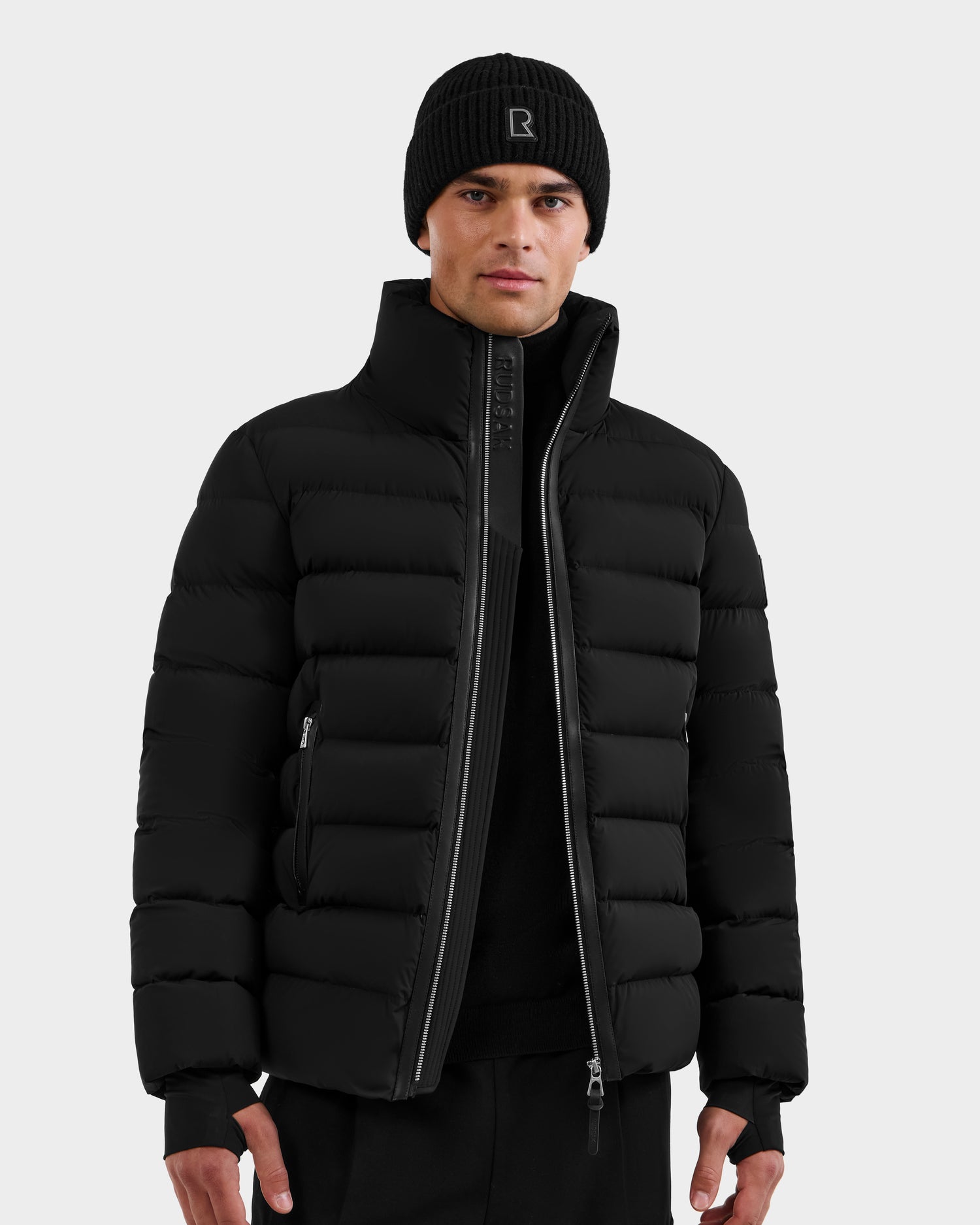 A person wearing the Rudsak NAVAN - 6125598 Black quilted down puffer, a black knit beanie, and black pants stands facing forward against a plain light gray background.