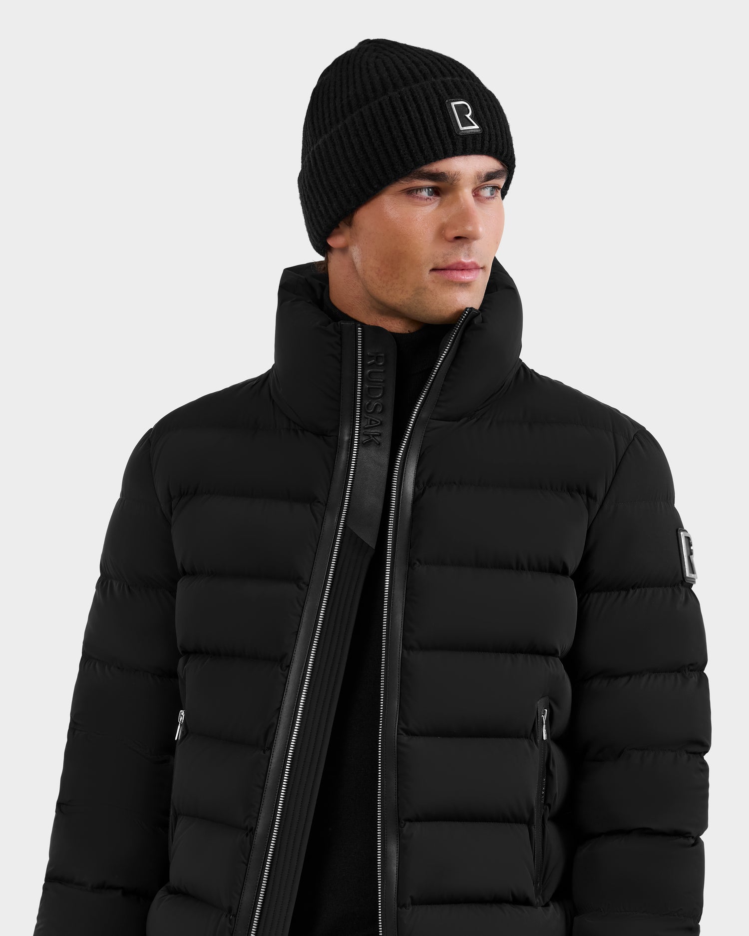 A man wears the Rudsak NAVAN - 6125598 Black quilted down puffer with a high collar and a black knit beanie with a small white logo, looking slightly left against a plain light background.