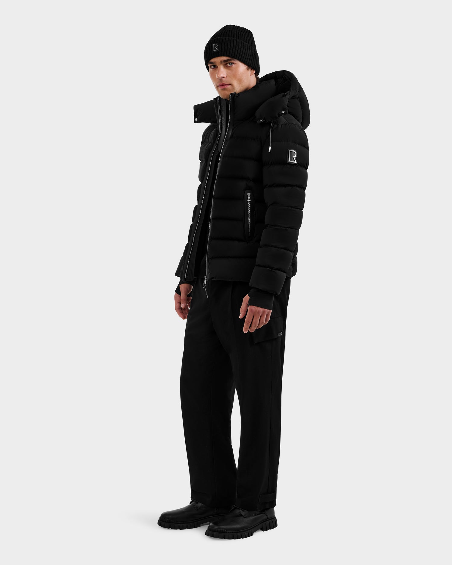 A person wearing the Rudsak NAVAN - 6125598 Black men’s quilted down puffer, paired with a black beanie, pants, and shoes, stands turned slightly to the side against a plain light background.