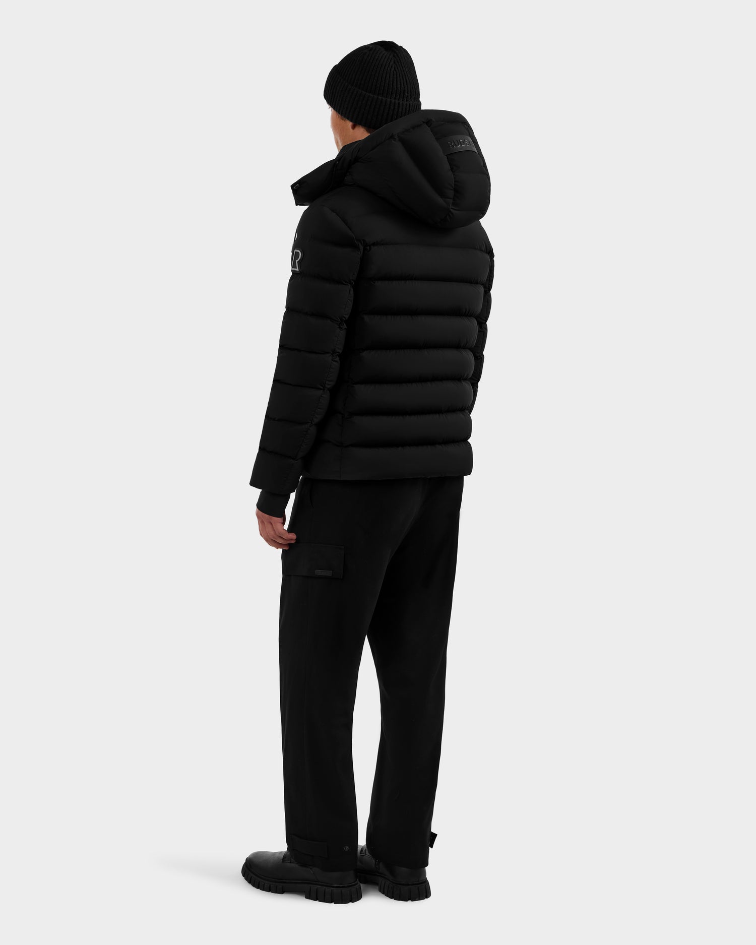 A person stands with their back turned, wearing the Rudsak NAVAN - 6125598 Black men's quilted down puffer, a black beanie, black pants with side pockets, and black shoes against a plain light background.