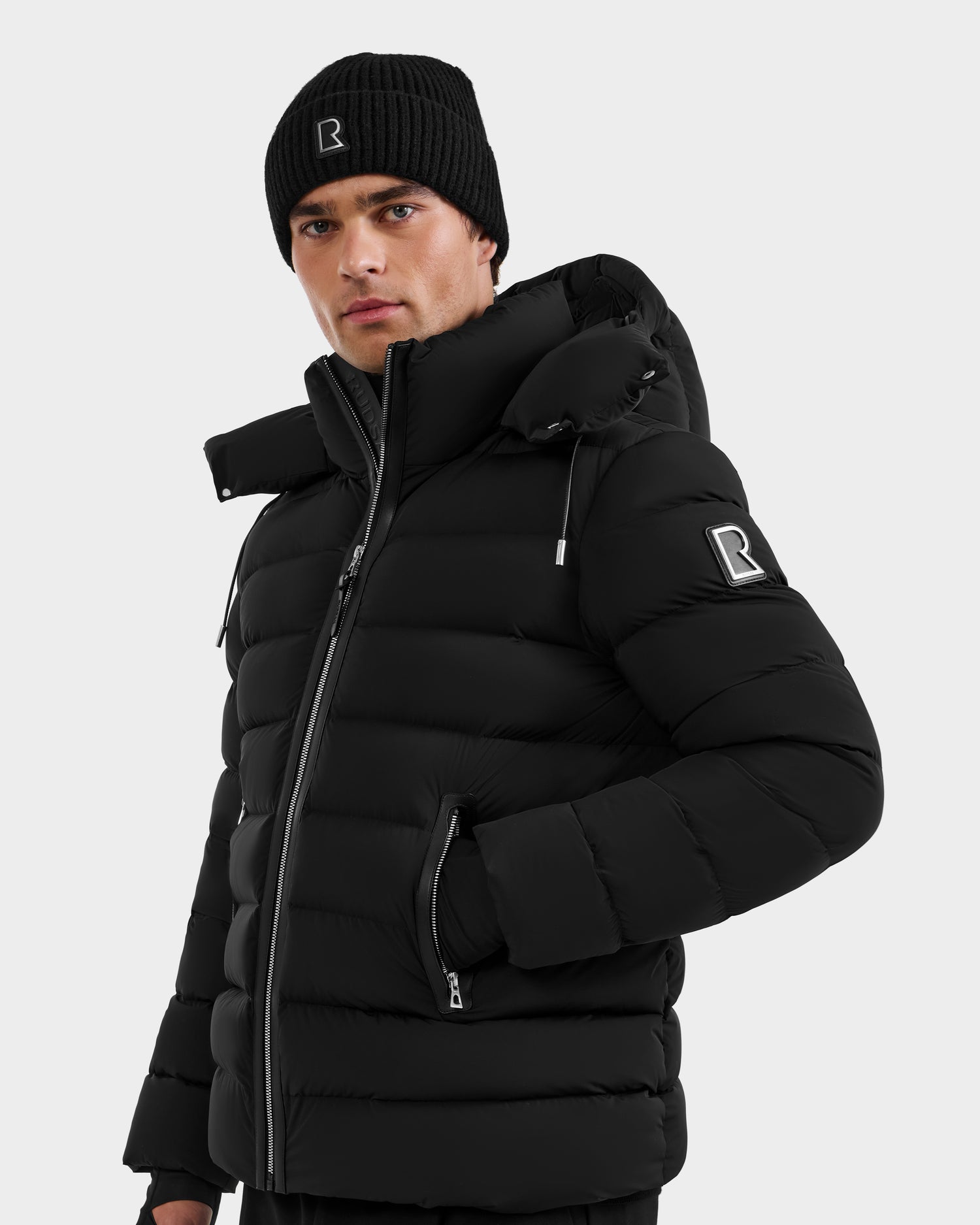 Wearing the Rudsak NAVAN - 6125598 Black men’s down puffer jacket with hood, zippered pockets, and high collar, plus a black beanie—both featuring a white R logo—a person stands against a light background.