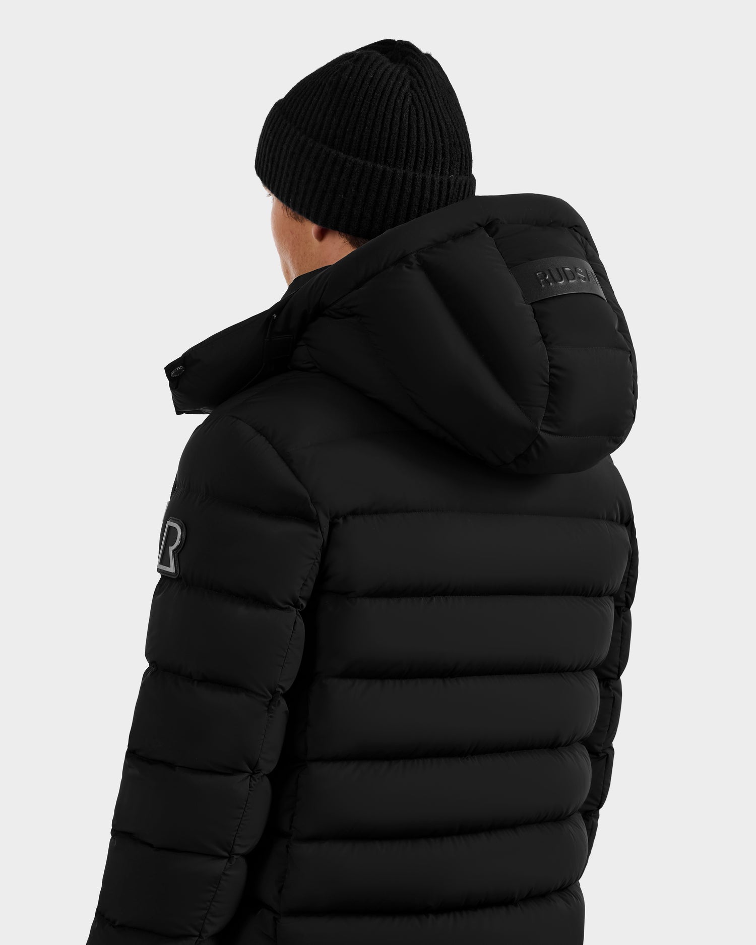 A person wearing the Rudsak NAVAN - 6125598 Black men's quilted down puffer jacket with a hood and a black knit beanie, shown from behind on a plain light background. The RDS-certified jacket has a sleeve patch and logo on the hood.