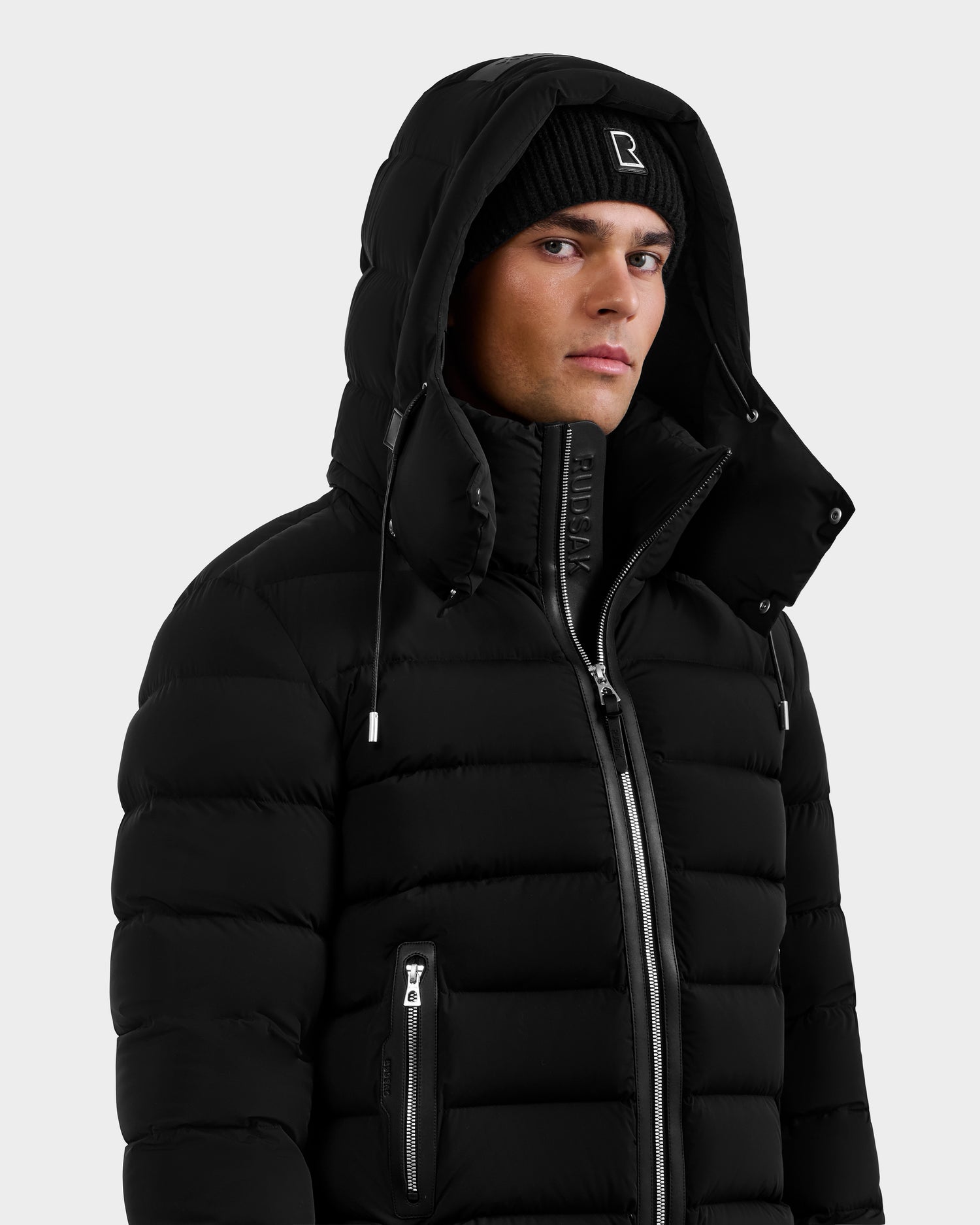 A man in the Rudsak NAVAN - 6125598 Black water-repellent quilted down puffer with hood up and a black knit beanie, featuring a visible zipper and zippered pockets, looks slightly to the side against a plain light background.
