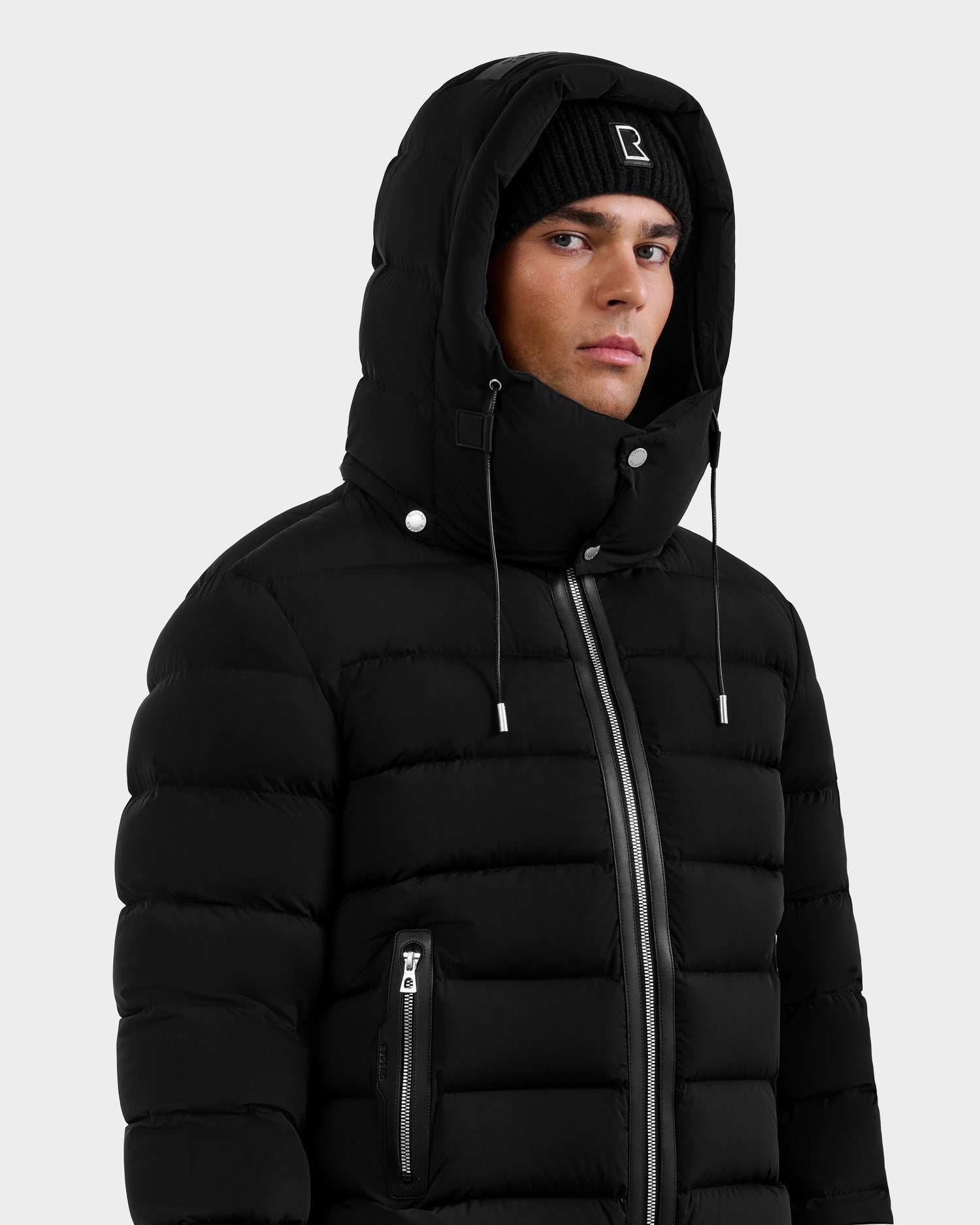 A model wears the Rudsak NAVAN - 6125598 Black men’s water-repellent quilted down puffer with hood up and silver zippers and buttons, paired with a black beanie against a plain light gray background.